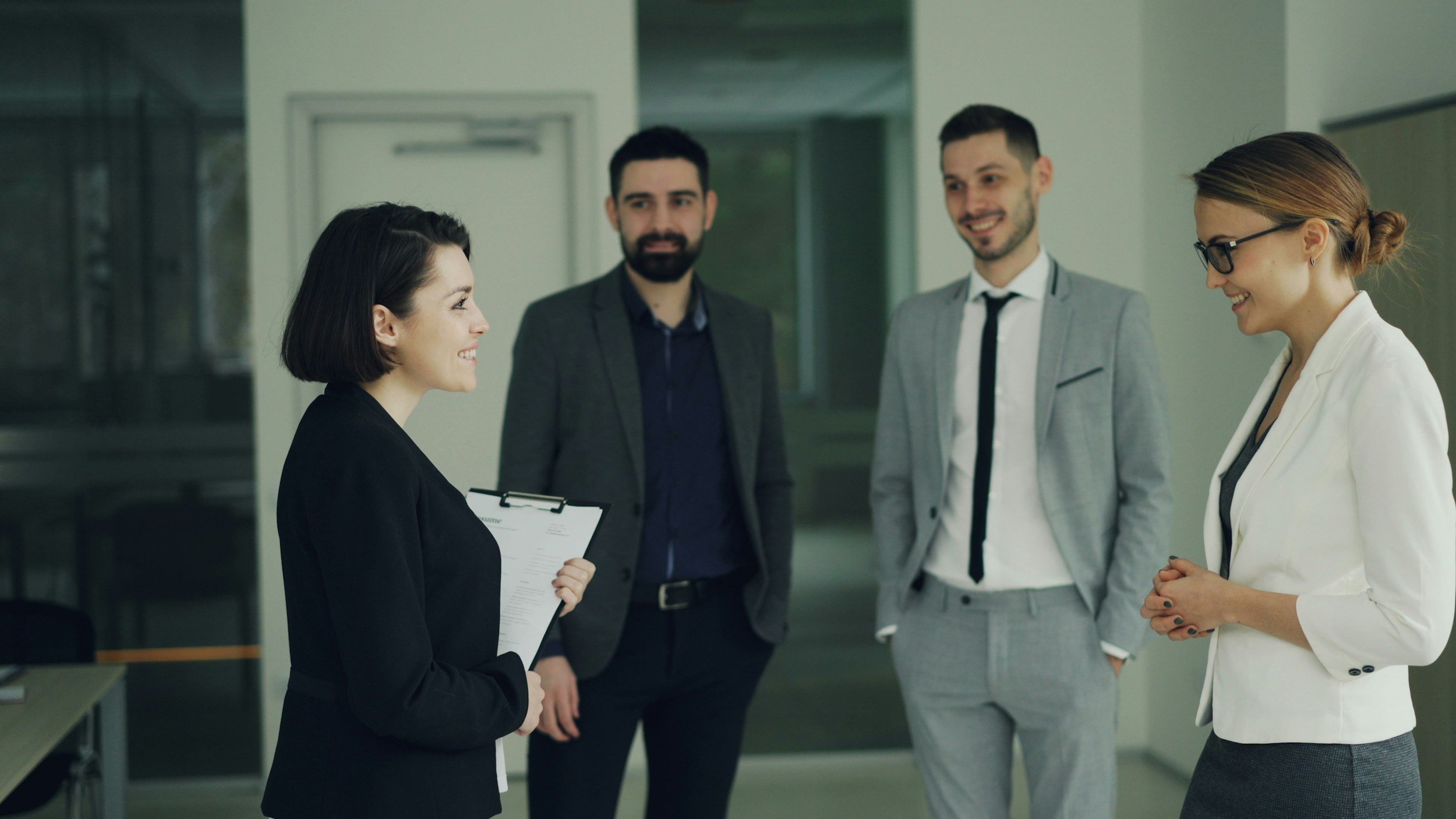 Four professionally dressed people talking and smiling in an office setting, with one woman holding a clipboard.