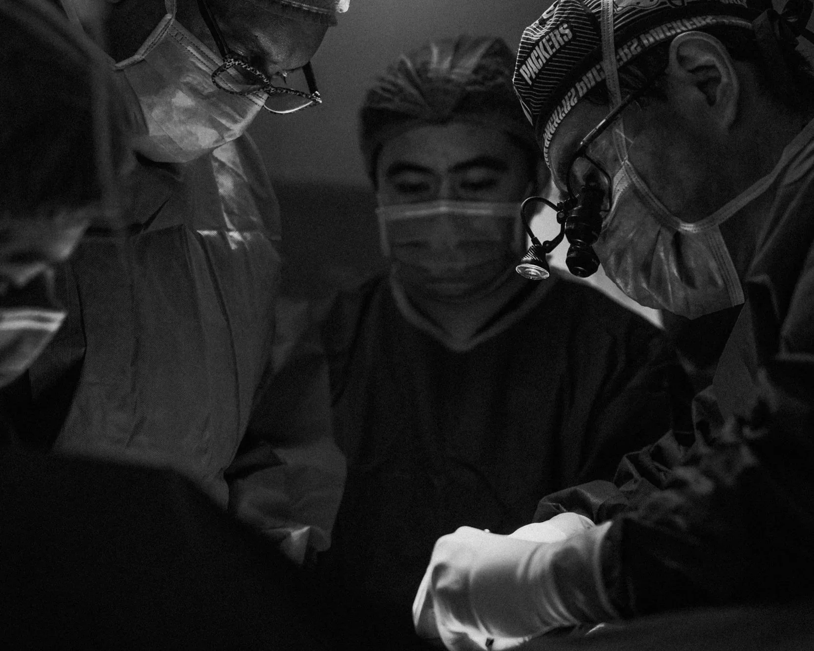 Three medical professionals, two men and one woman, wearing surgical scrubs, masks, and headlamps, are gathered closely around a patient, performing a medical procedure in a dimly lit environment.