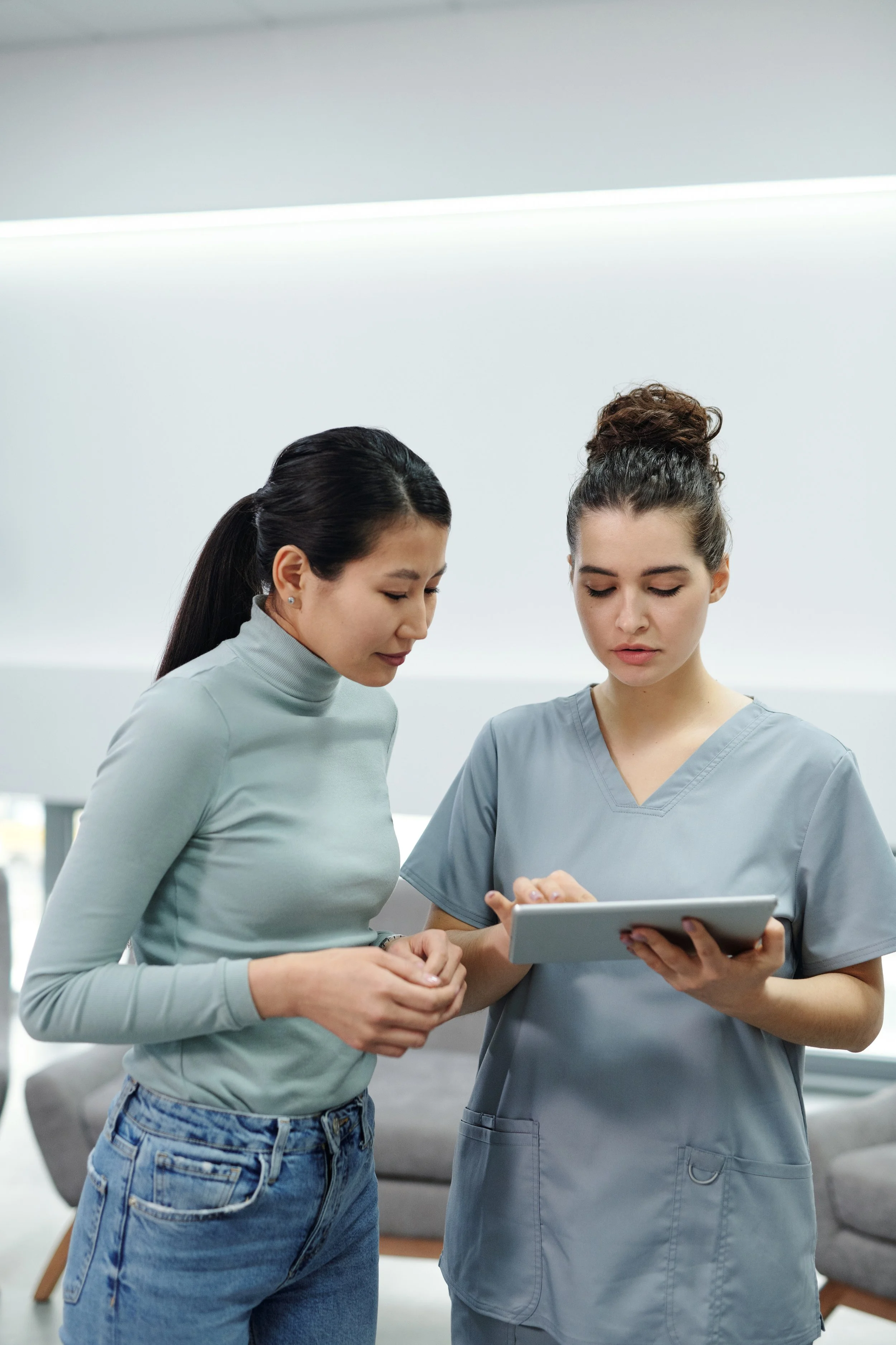 Two women, one in casual clothing and the other in scrubs, are looking at a tablet together in a modern indoor setting.