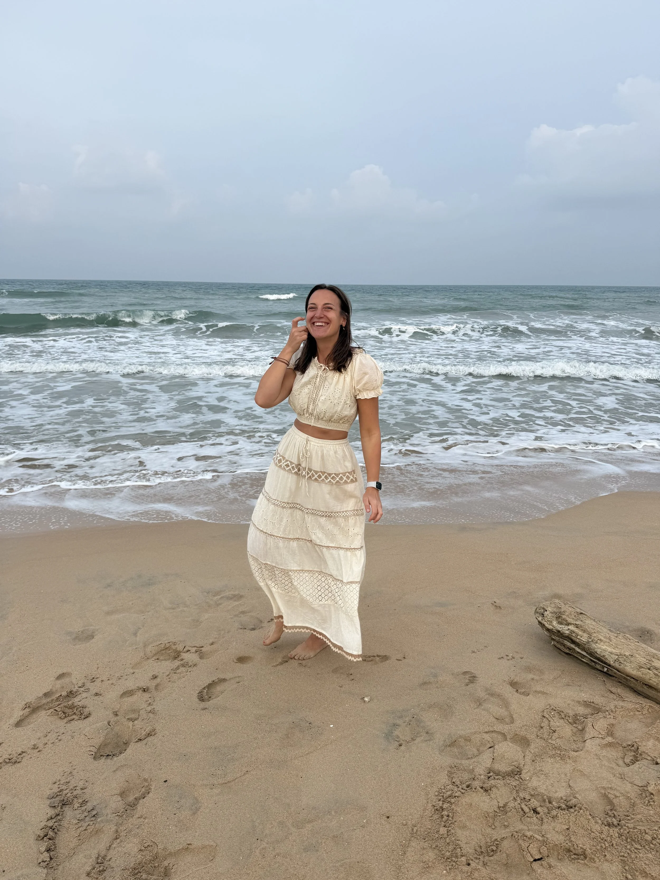 A woman smiling and standing barefoot on the sandy beach near the ocean with waves and cloudy sky in the background, wearing a cream-colored, crochet dress.