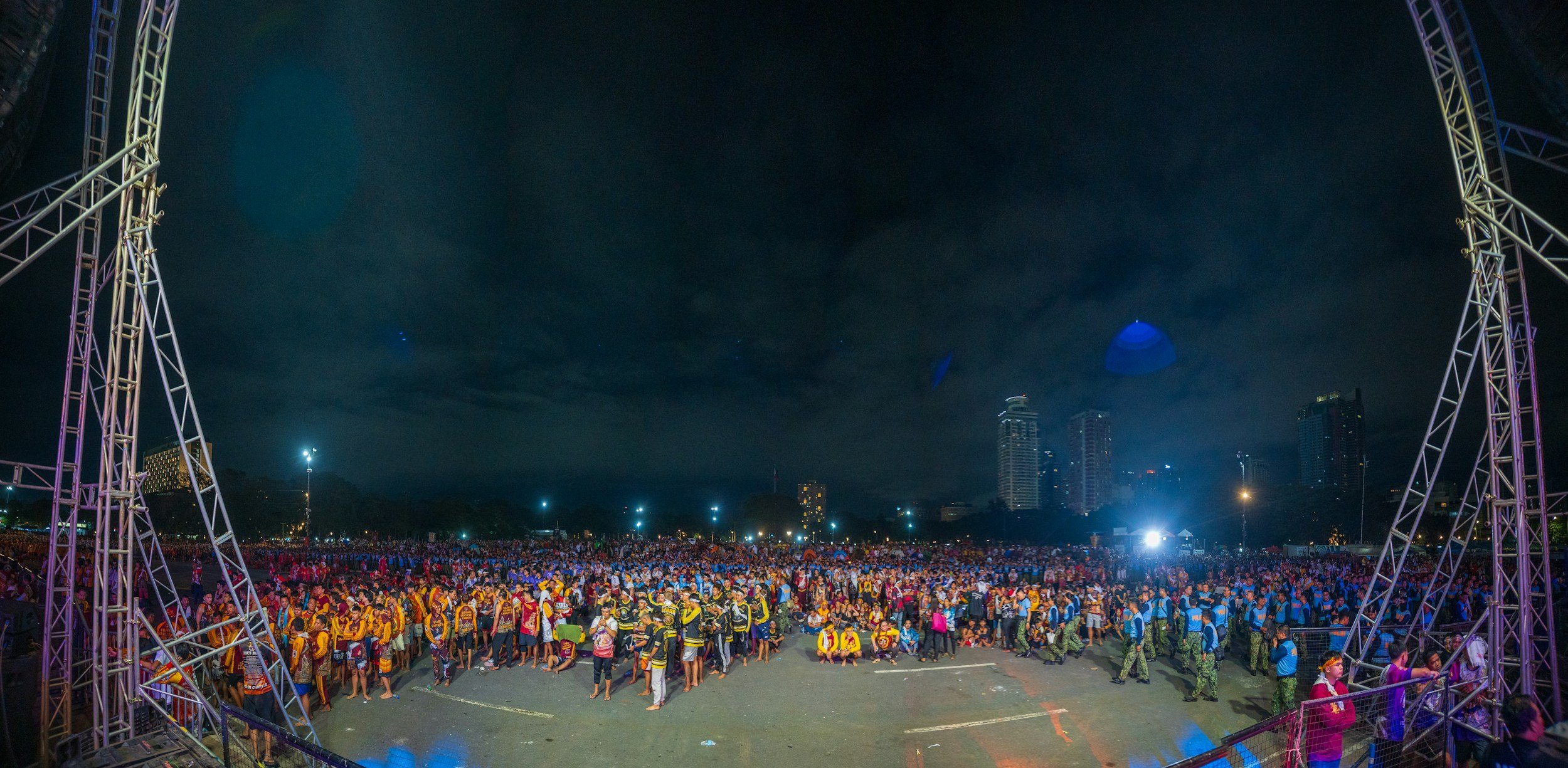 A large crowd of people at a nighttime outdoor event under a dark sky, with city buildings in the background and a stage framed by metal scaffolding.