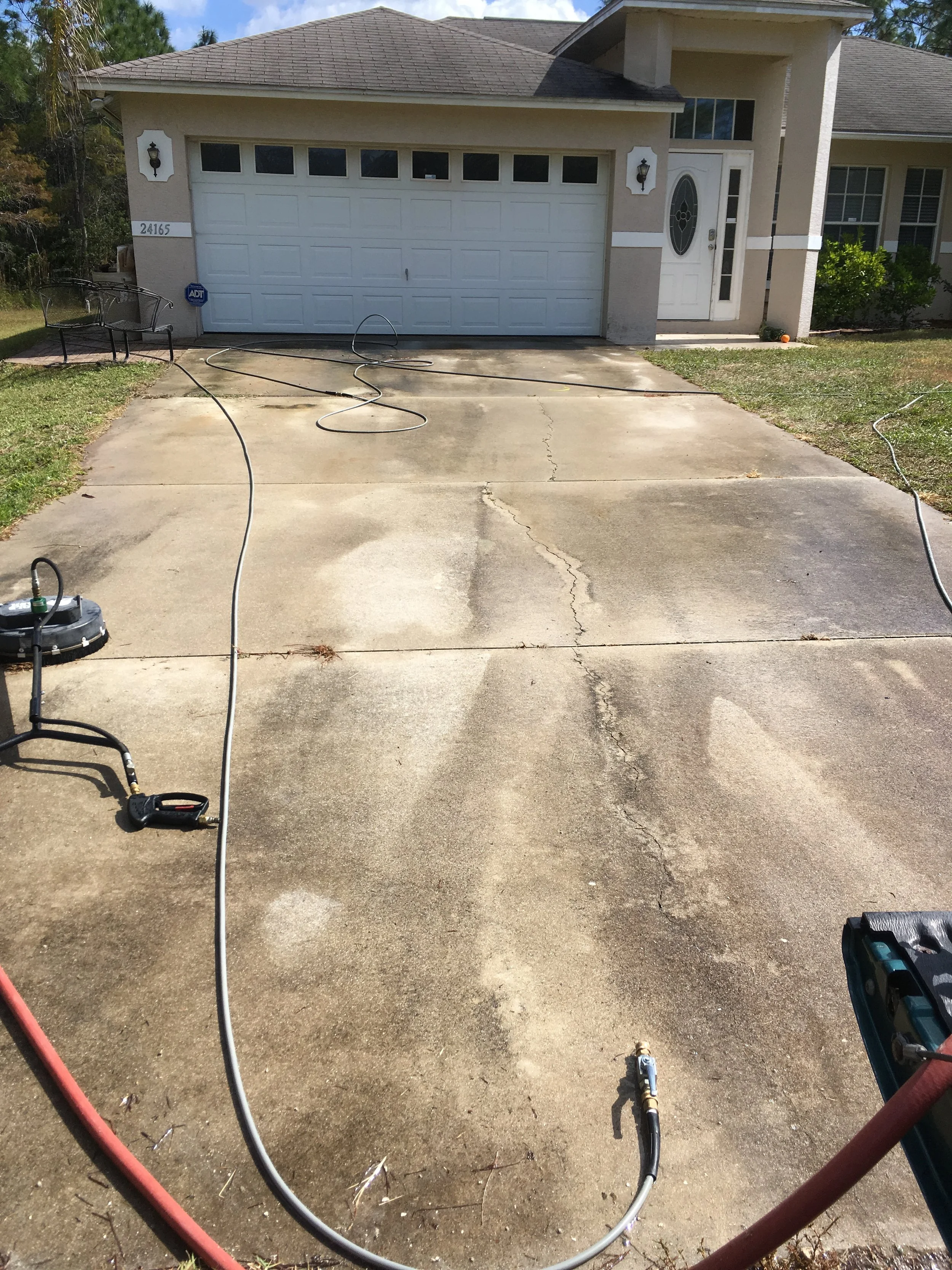 A residential driveway with some gardening or cleaning equipment, including a hose, a pressure washer, and some nozzles, nearby. The house has a garage with a white door, and the house has beige walls with white accents. The lawn has some shrubs and a pumpkin on the right side.