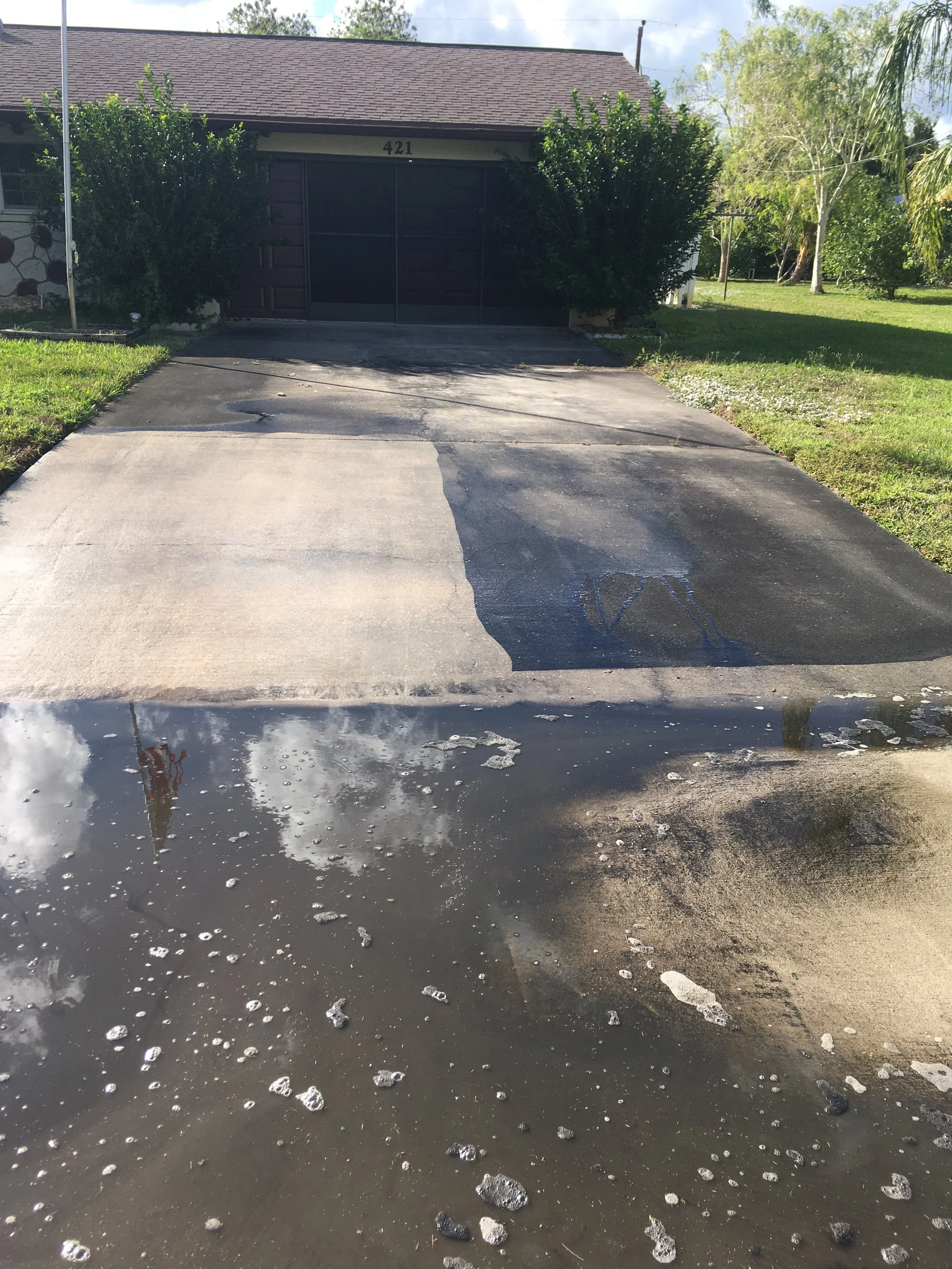 A driveway with water and soap suds, leading to a house with a garage and bushes on the sides. The house has a brown roof and one visible house number 421.
