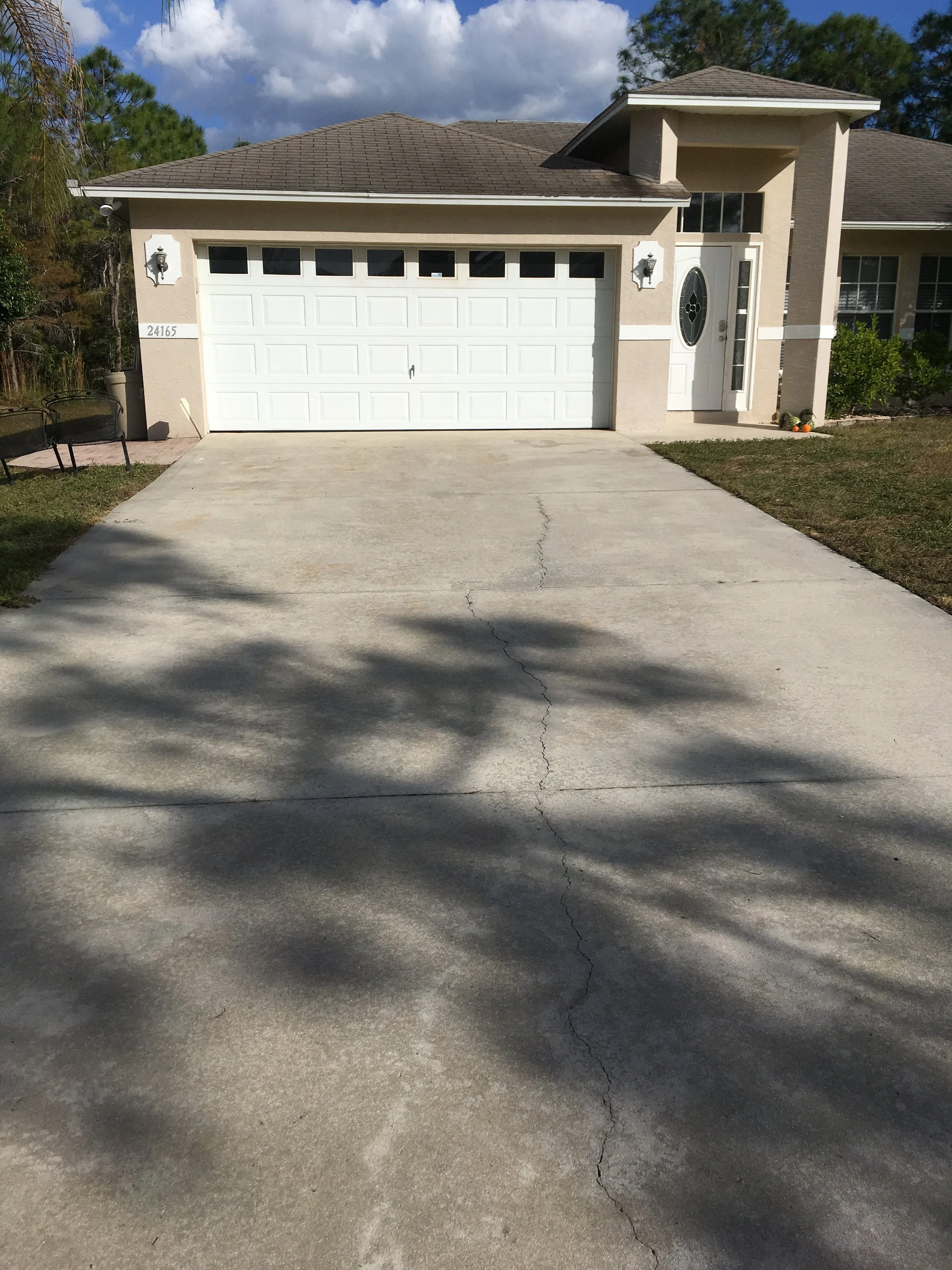 Front view of a beige house with a white garage door, two outdoor light fixtures, and a driveway with a visible crack. Small porch area with a white front door and decorative glass window, lawn, and trees in the background.