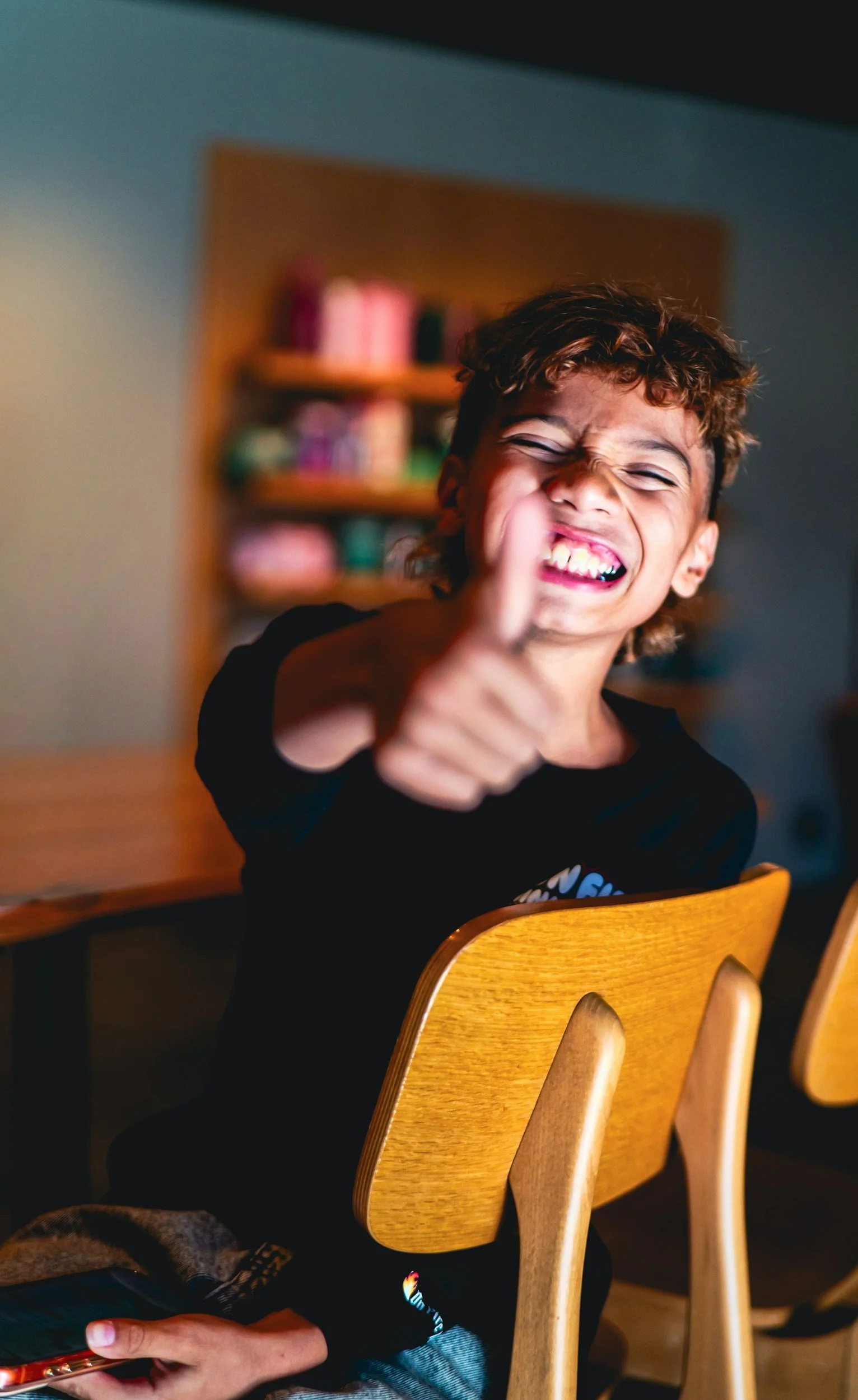 A young boy with curly brown hair and a black shirt is smiling broadly with his eyes closed, showing his teeth. He is sitting at a wooden dining chair and pointing towards the camera with his finger. In the background, there is a blurred bookshelf with colorful books, and part of another person's hand holding a phone is visible at the bottom of the image.