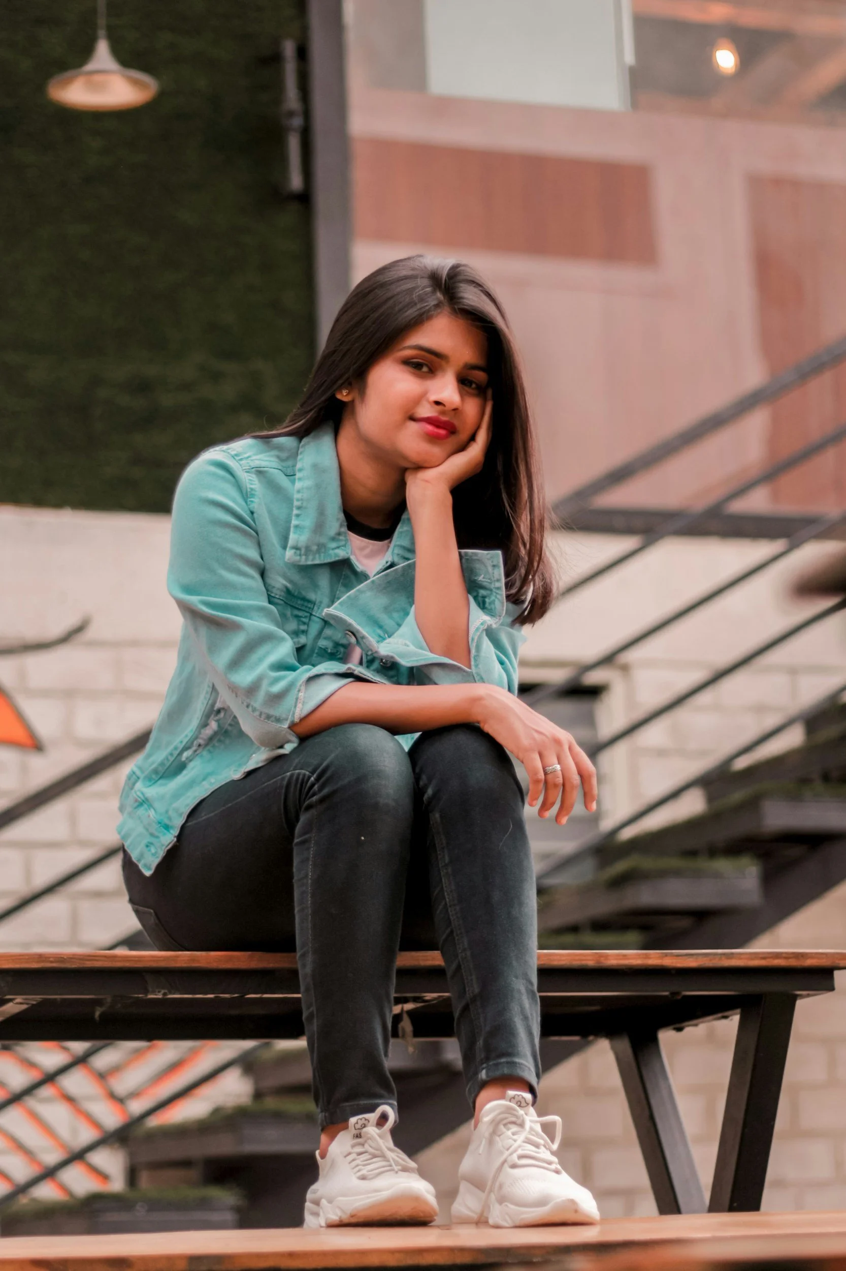 A young woman sitting on outdoor stairs, resting her chin on her hand, wearing a light blue jacket, black jeans, and white sneakers, with a background of a modern building and a staircase railing.