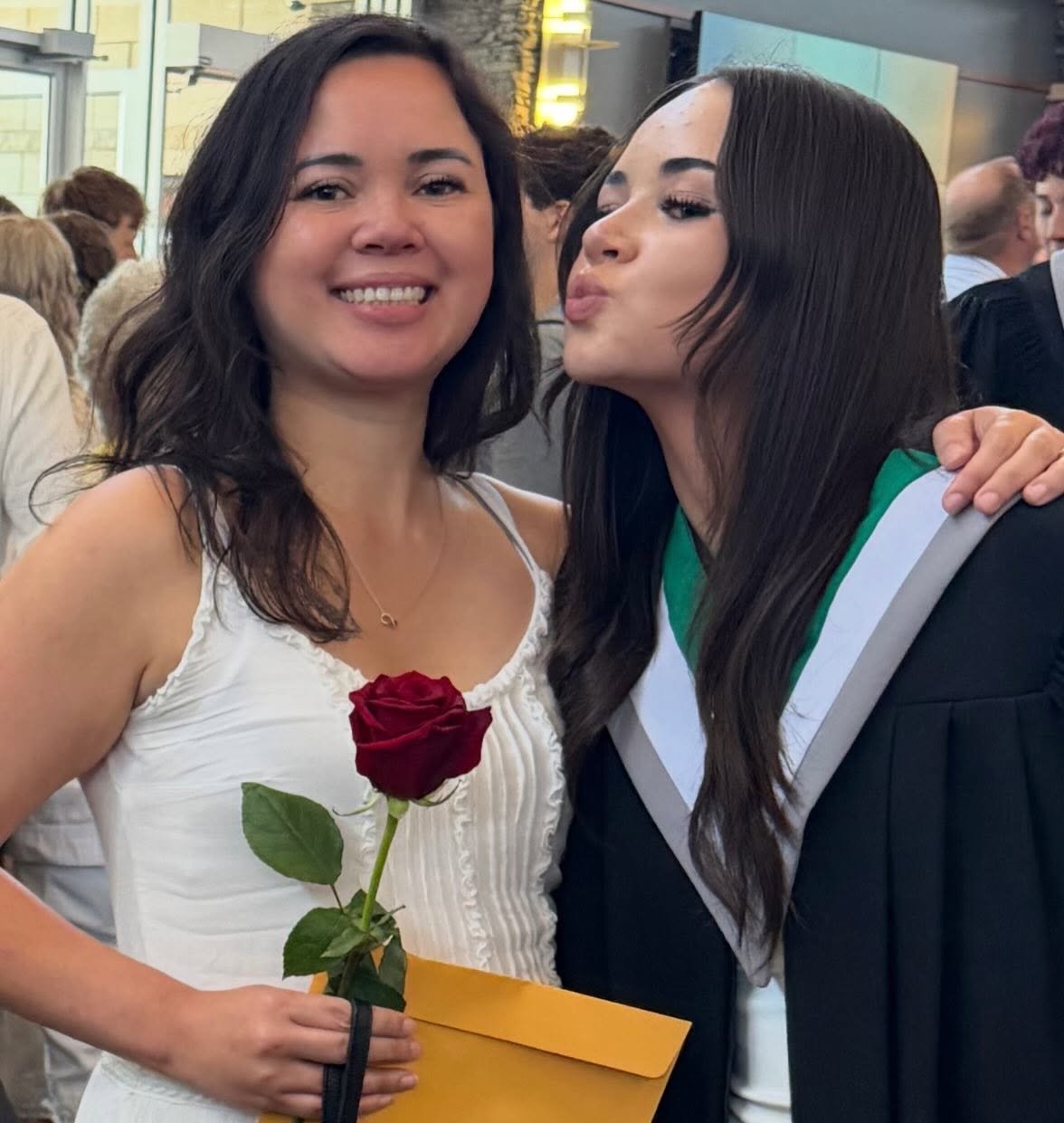A woman in a white dress holding a red rose and an envelope, smiling next to a woman in a graduation gown giving a kiss on the cheek.
