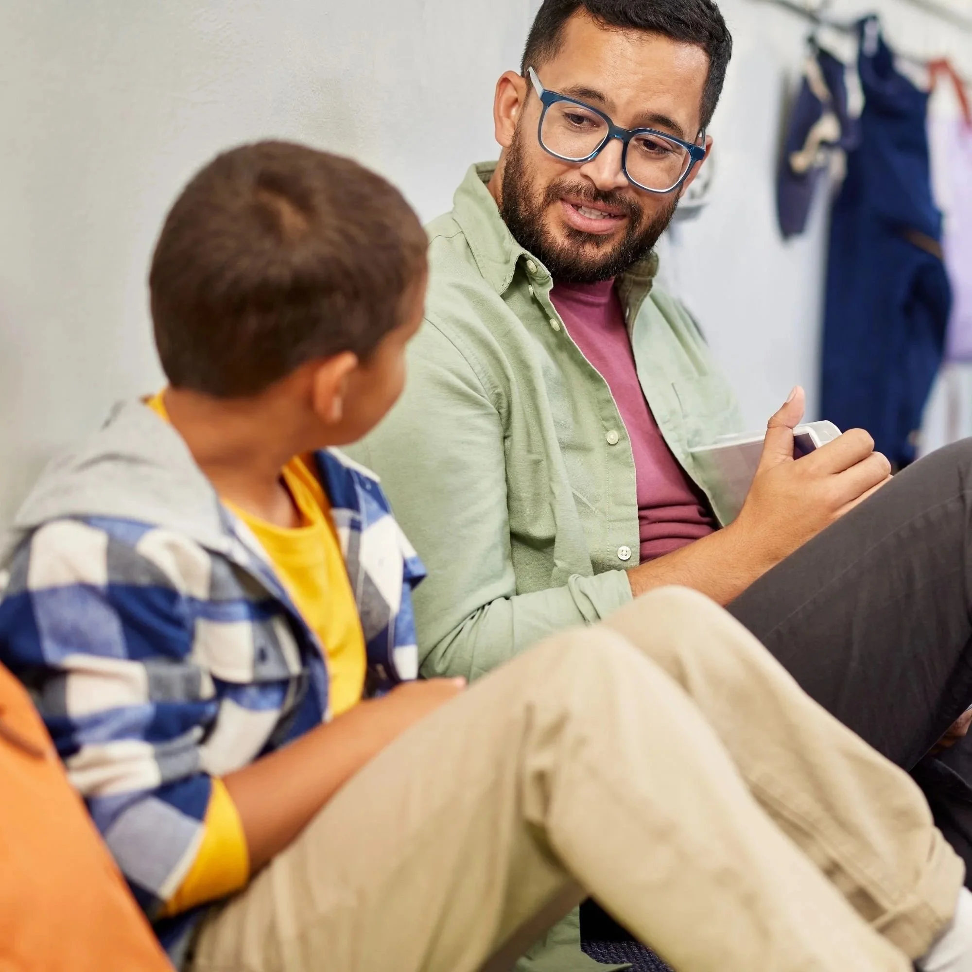 A man with dark hair, glasses, and a beard, wearing a green shirt and pink T-shirt, is talking to a child with short brown hair, who is wearing a striped blue and white shirt with yellow accents, in a classroom or similar setting.