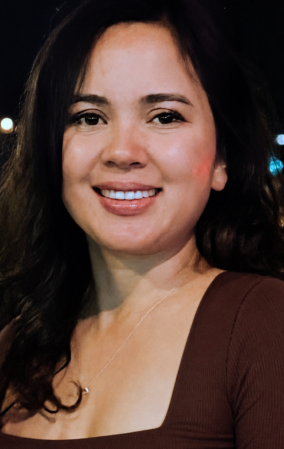 A woman with long, dark, wavy hair smiling at night, wearing a brown top and a necklace with a small pendant.