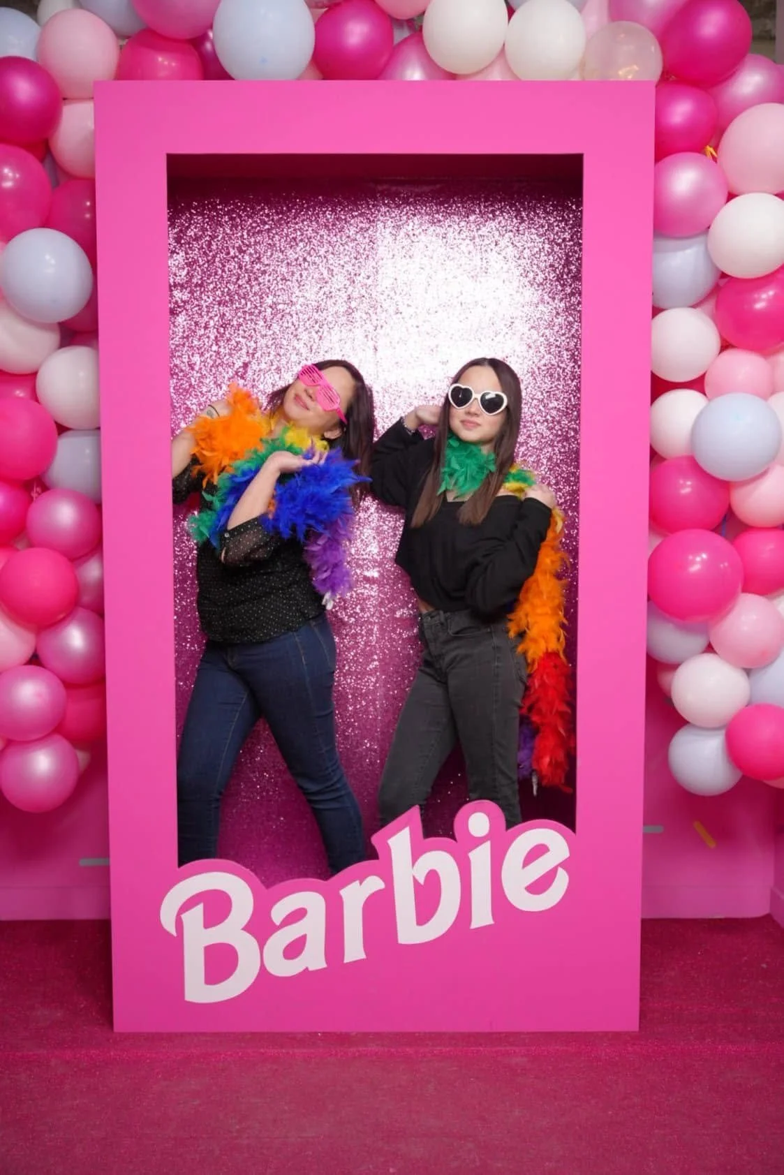 Two women posing inside a pink photo frame with sparkly background, surrounded by pink and white balloons, wearing colorful feather boas and novelty sunglasses, at a Barbie-themed event.