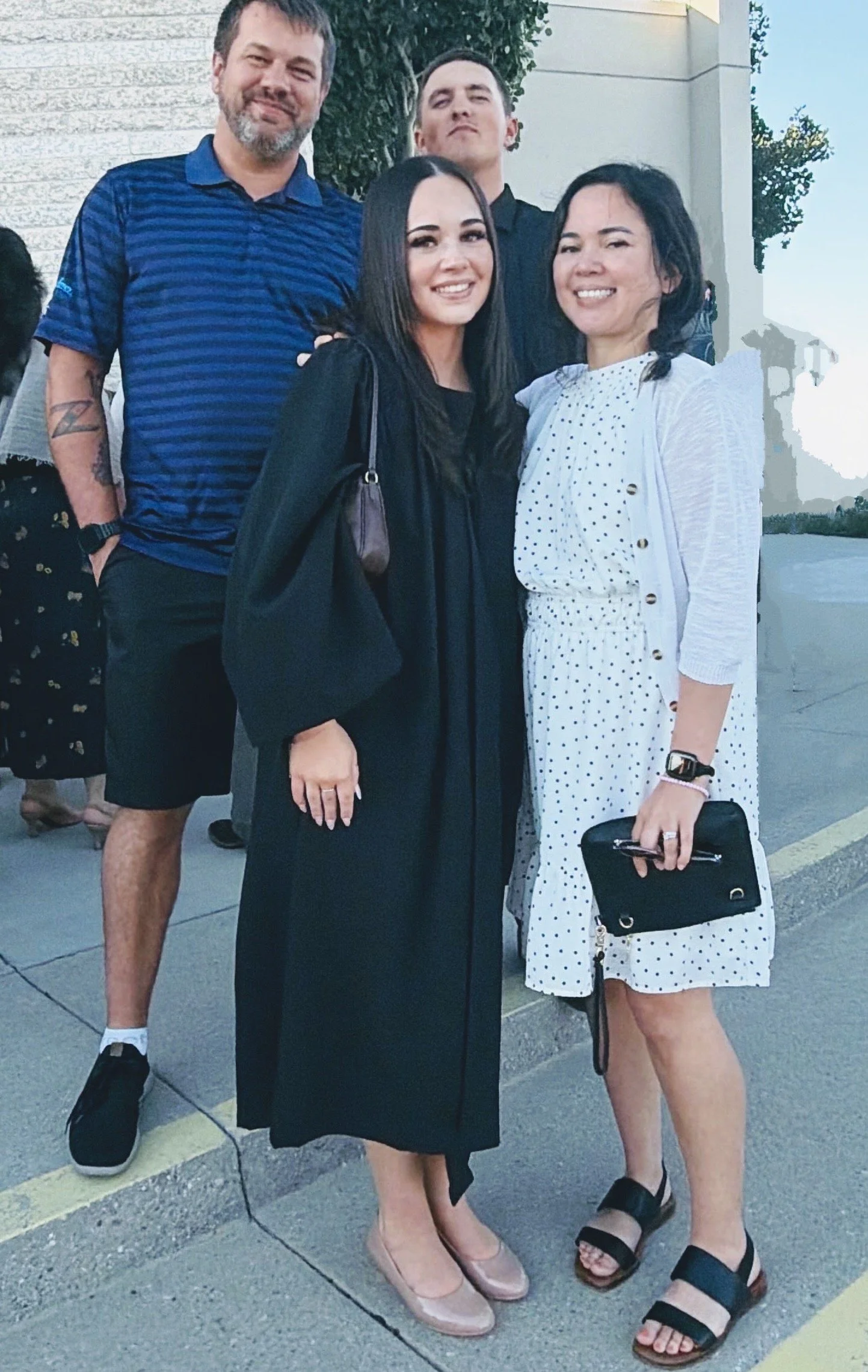 Four people standing outside on a sidewalk smiling and posing for a photo, with a building and trees in the background.