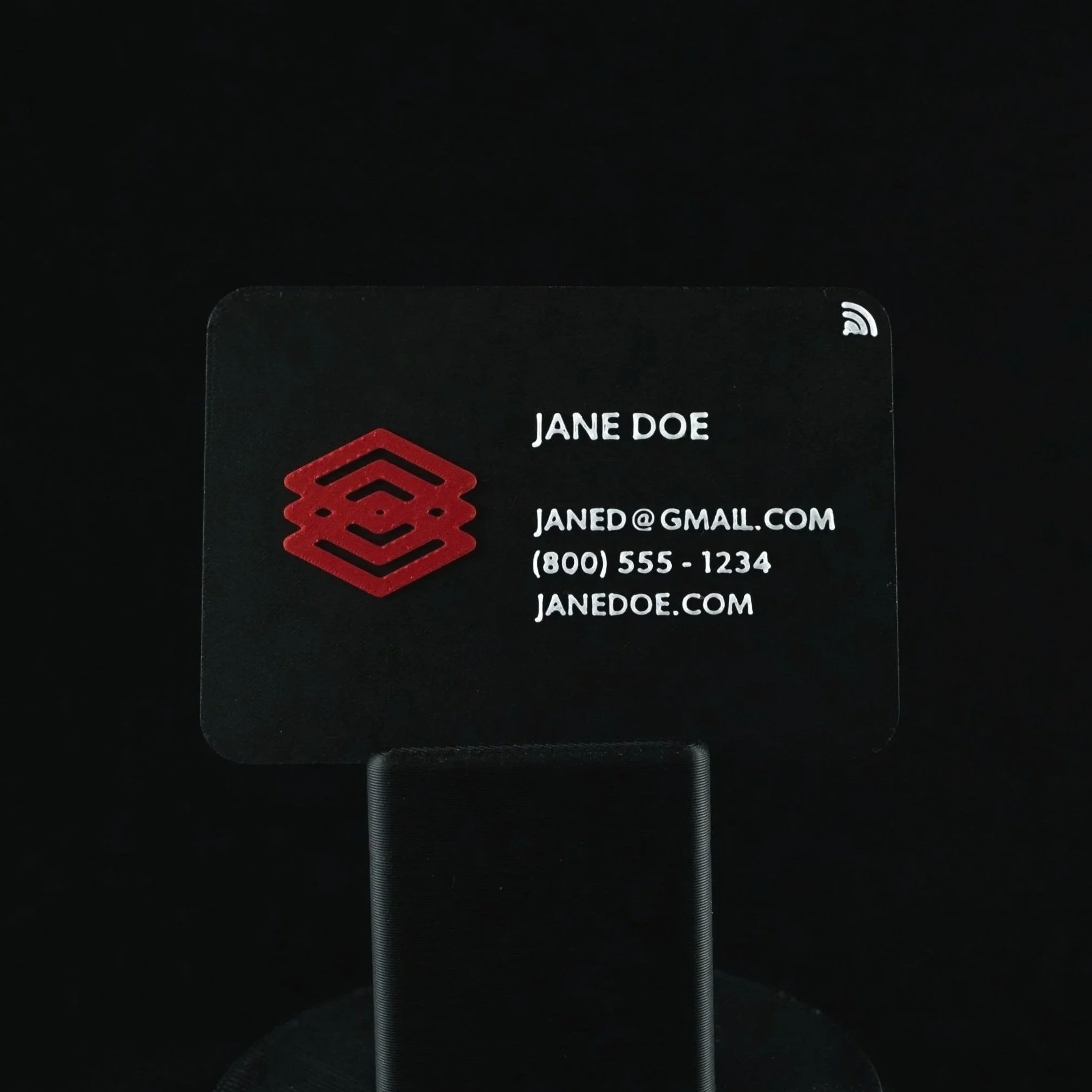 Close-up of a black 3D-printed business card with white text and a red geometric logo, showing an email address and phone number.