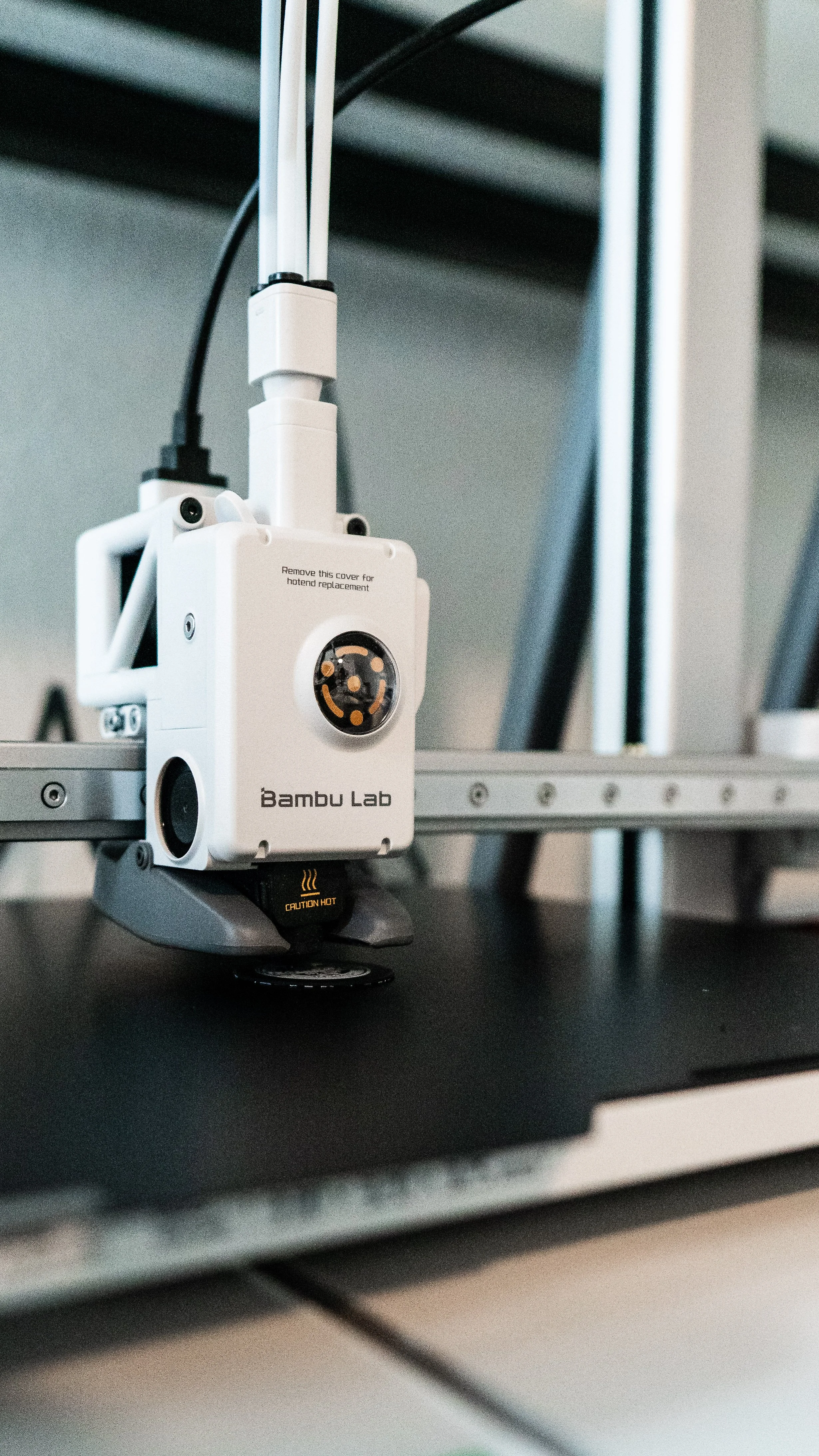 A white 3D printer labeled 'Bambu Lab' is on a dark surface with a yellow 'Caution Hot' indicator below it. The printer is connected to power and placed near a metal frame.