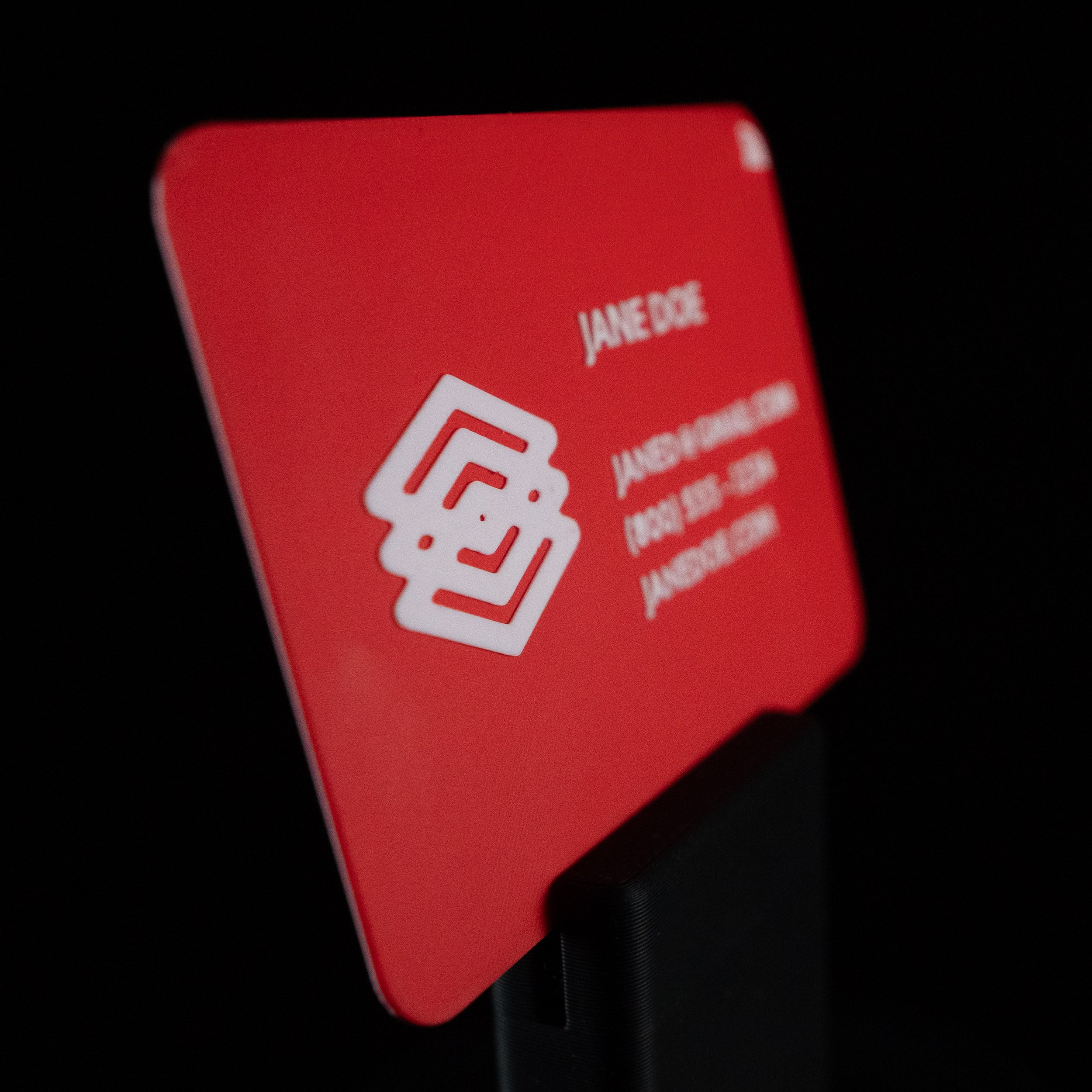 Close-up of a red 3D-printed NFC business card with white text and a red geometric logo, showing an email address and phone number.