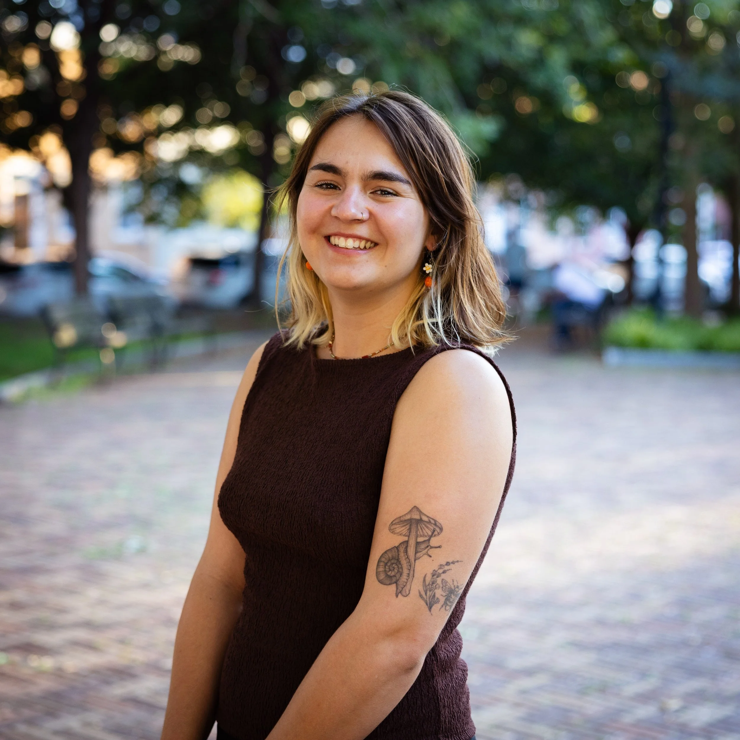 Co-founder Ayanna with brown hair and tattoos smiling outdoors in a park with trees in the background.