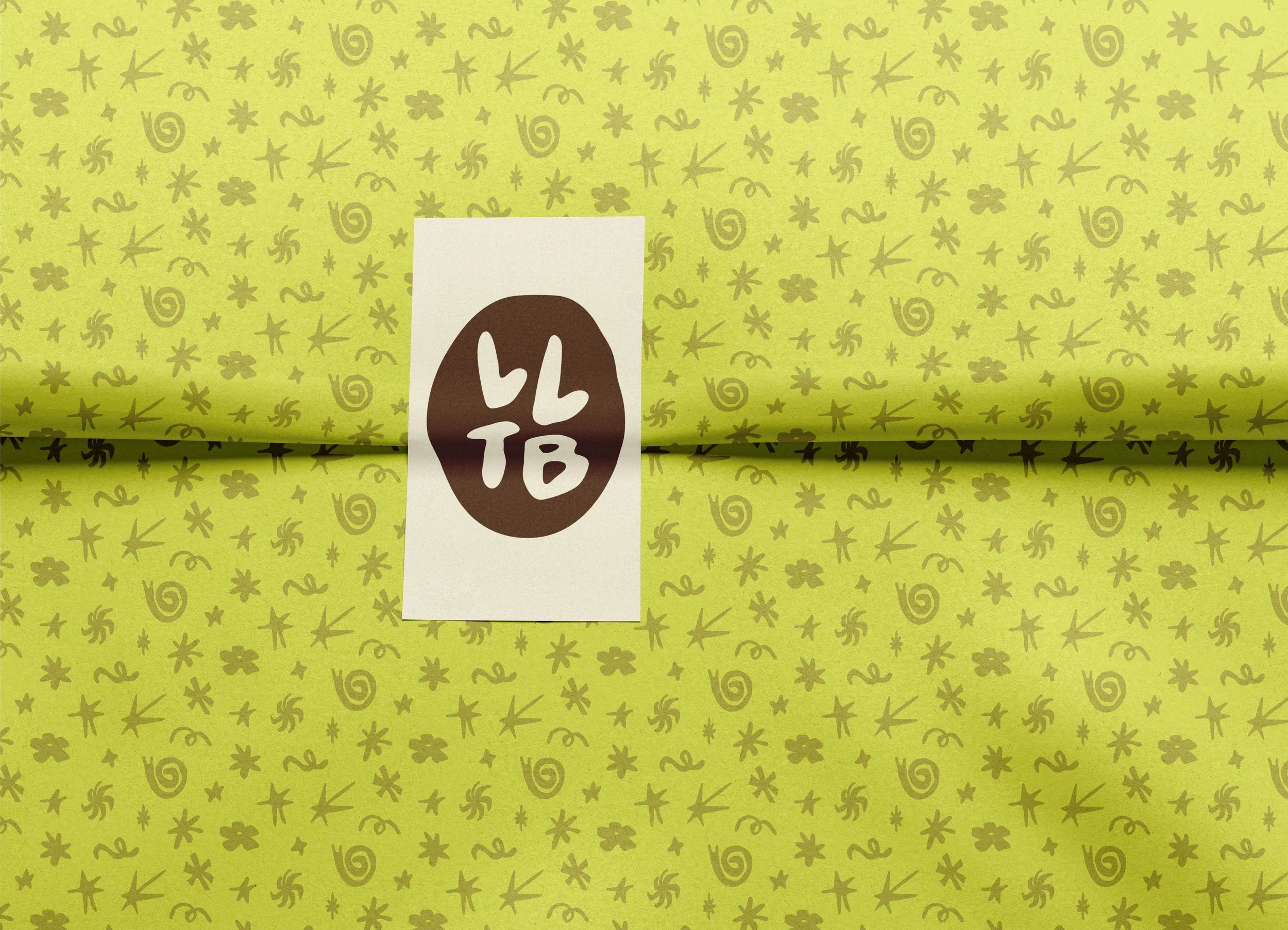 Green wrapping paper with a pattern of various small doodles like stars, spirals, and abstract shapes, partially covering a white piece of paper with a brown circular sticker that reads "LLTB."