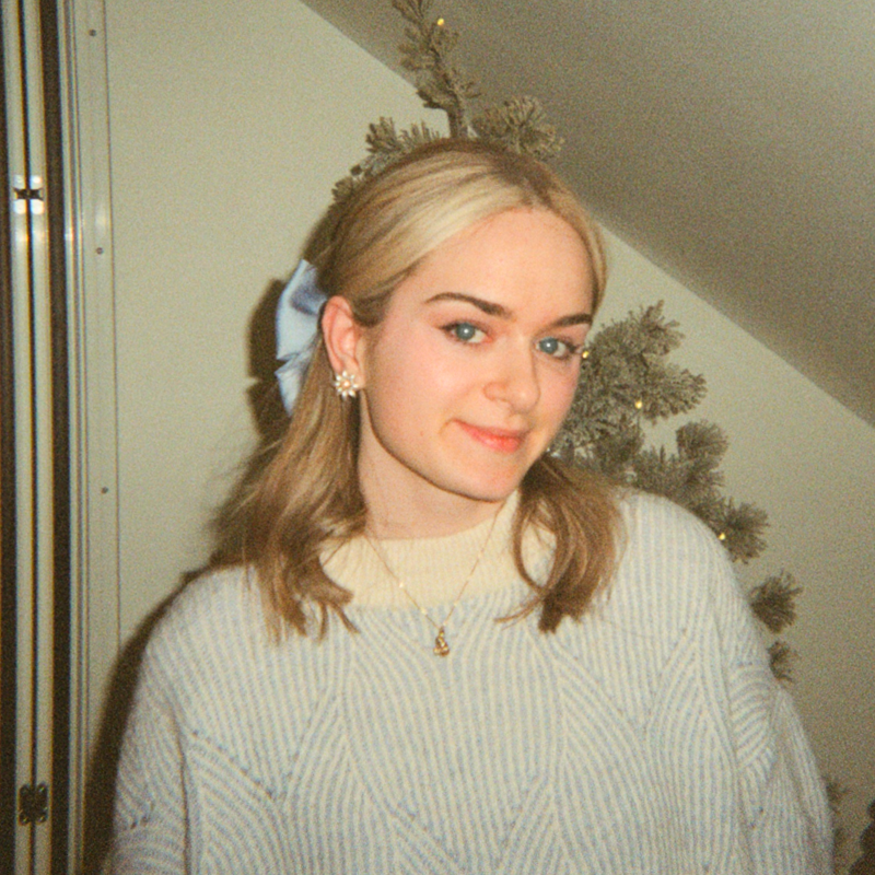 A young woman, called Chloe Daniels, with blonde hair wearing a cream sweater, gold necklace, and floral earrings, standing in front of a Christmas tree with soft lighting. She is the Founder of Frontier Marketing.