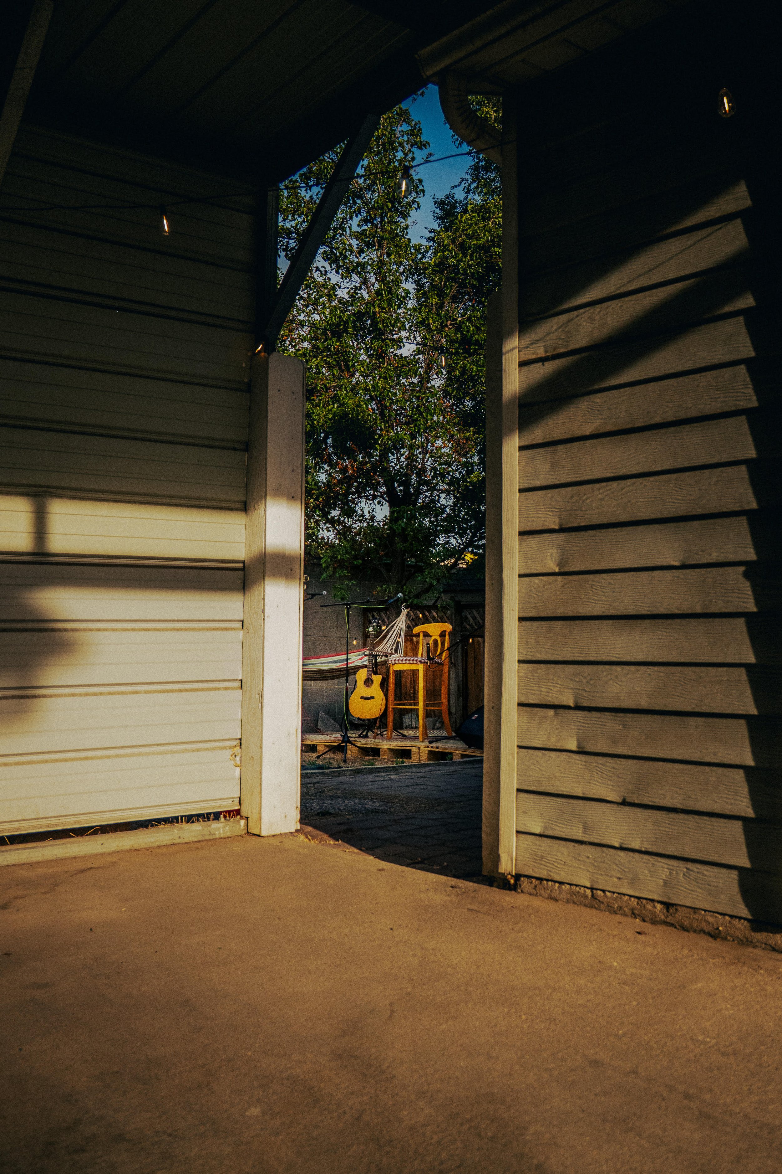 View from inside a partially open garage showing a yard with a guitar and a chair, trees in the background, and a blue sky.