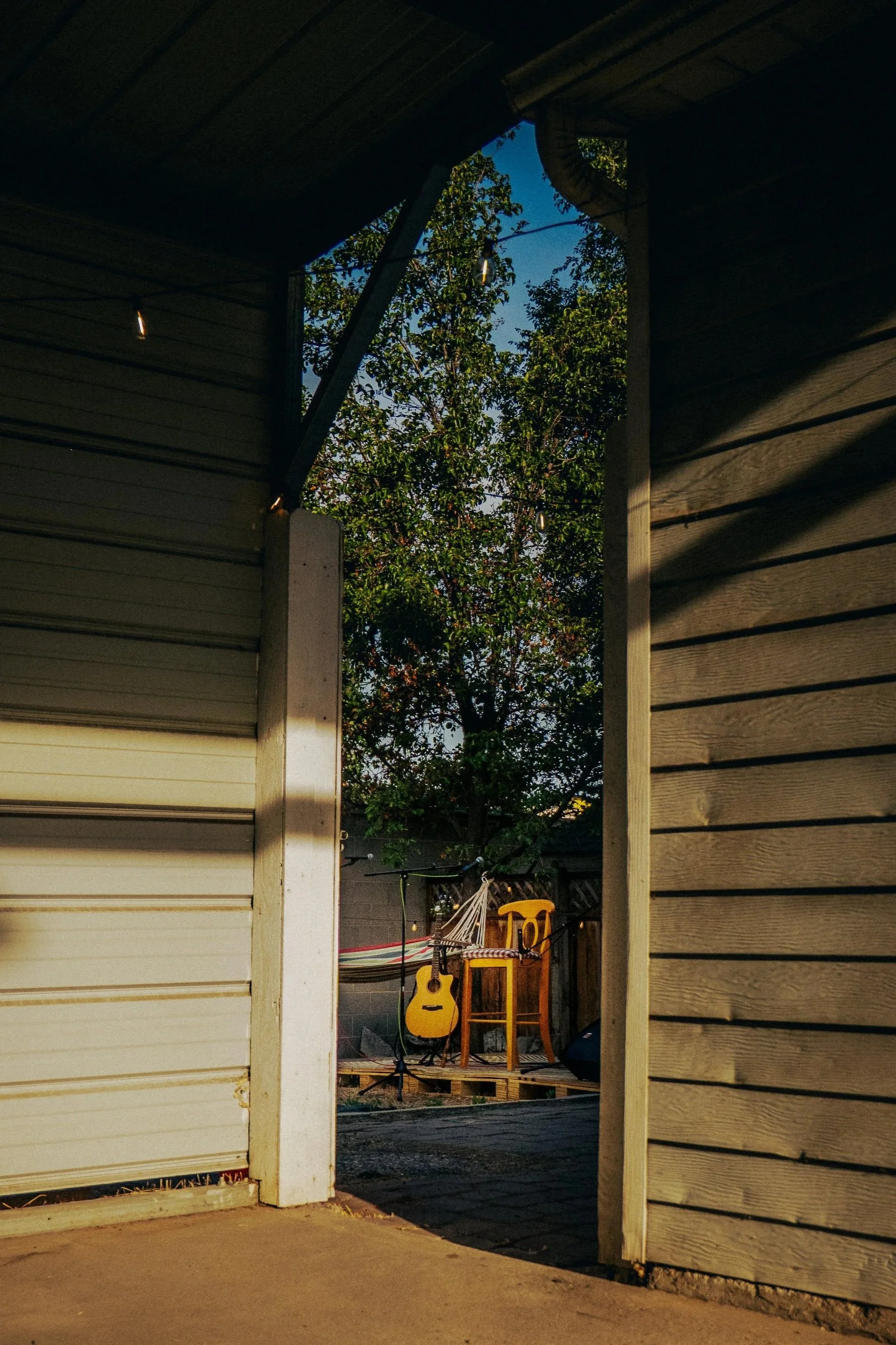 View of a backyard with a guitar, chair, and hammock seen through a partially open garage or shed door, with trees in the background and evening light.