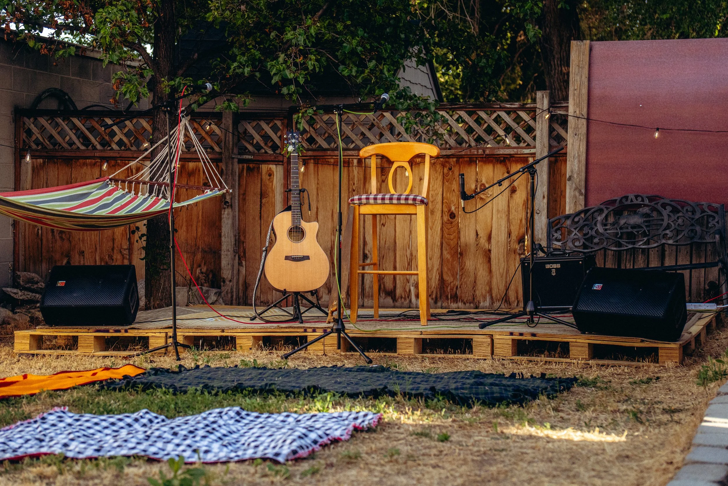 Outdoor stage set up with a guitar, stool, and microphone in front of a wooden fence, with string lights overhead and a hammock to the left.