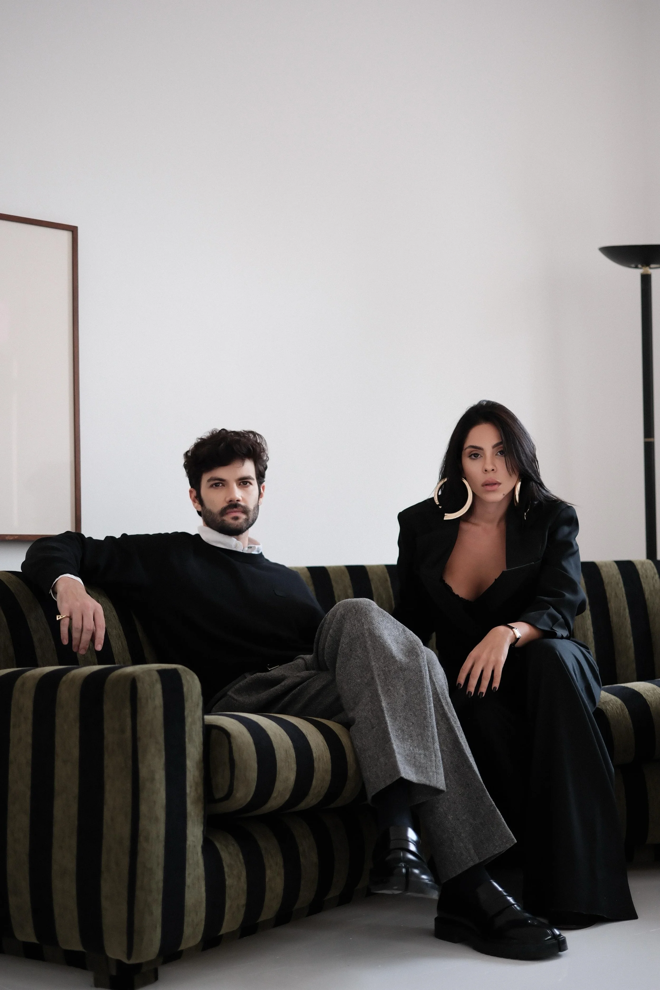 Carla Cruz e Philipe Pinheiro, do Estudio Sala, sentados em um sofá listrado em preto e bege, em ambiente de interiores minimalistas.