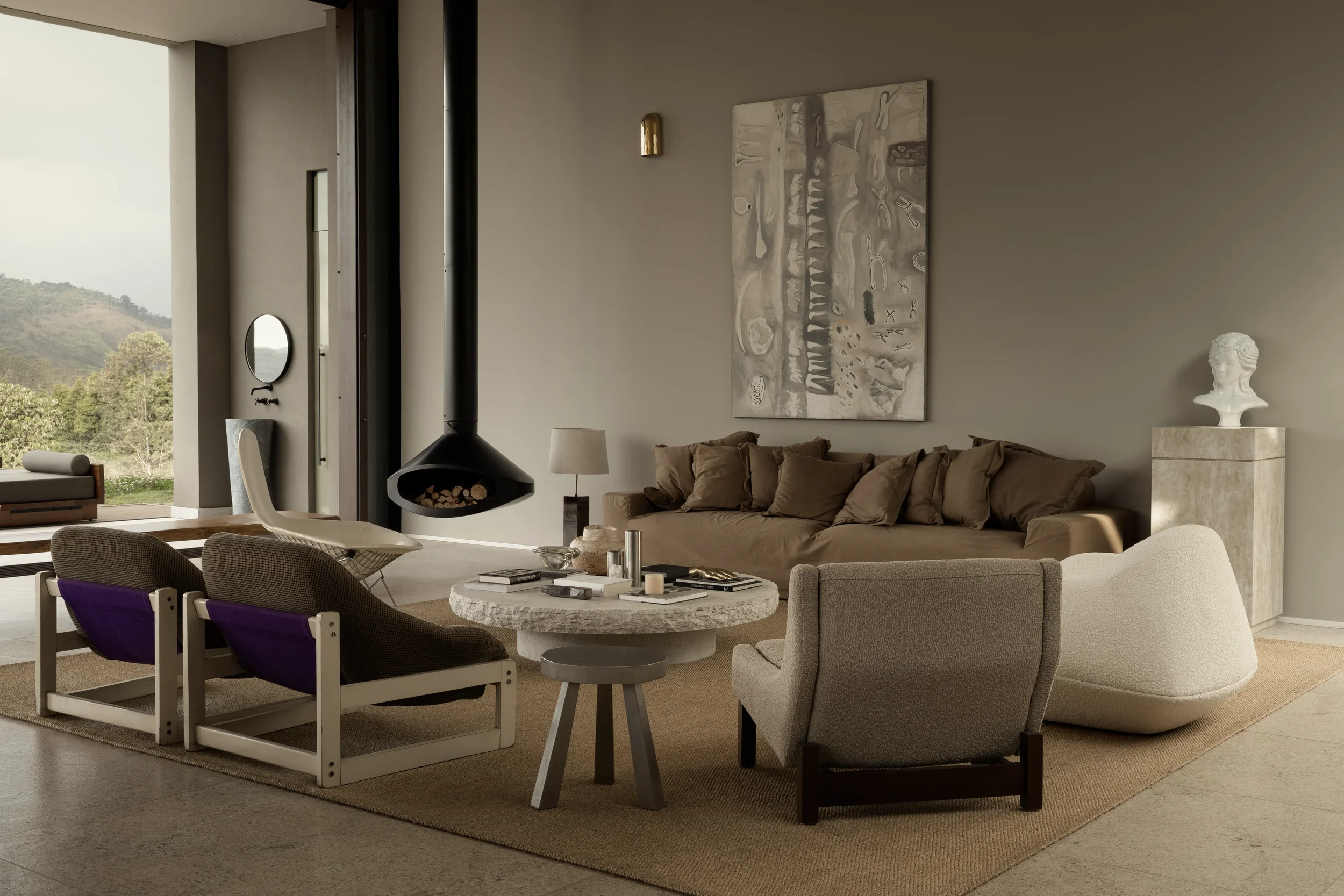 Modern living room with beige sofas, a coffee table, a white armchair, abstract wall art, and a fireplace with logs. Large window shows outdoor landscape with hills and trees.