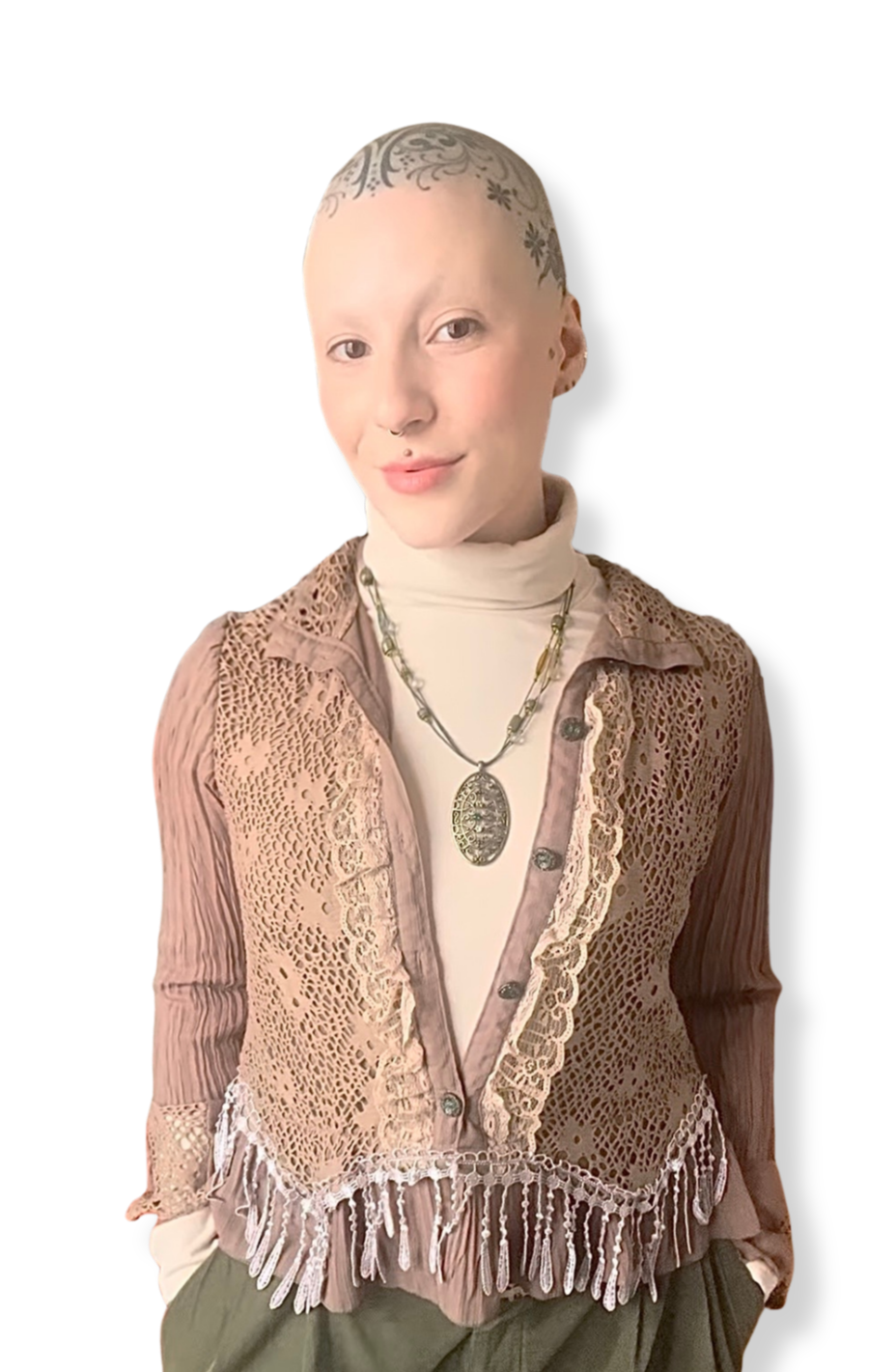 Yvi, a white, bald, agender person with an ornamental head tattoo, wearing a cream turtleneck, tan fringed button up blouse, green slacks, and an oval necklace
