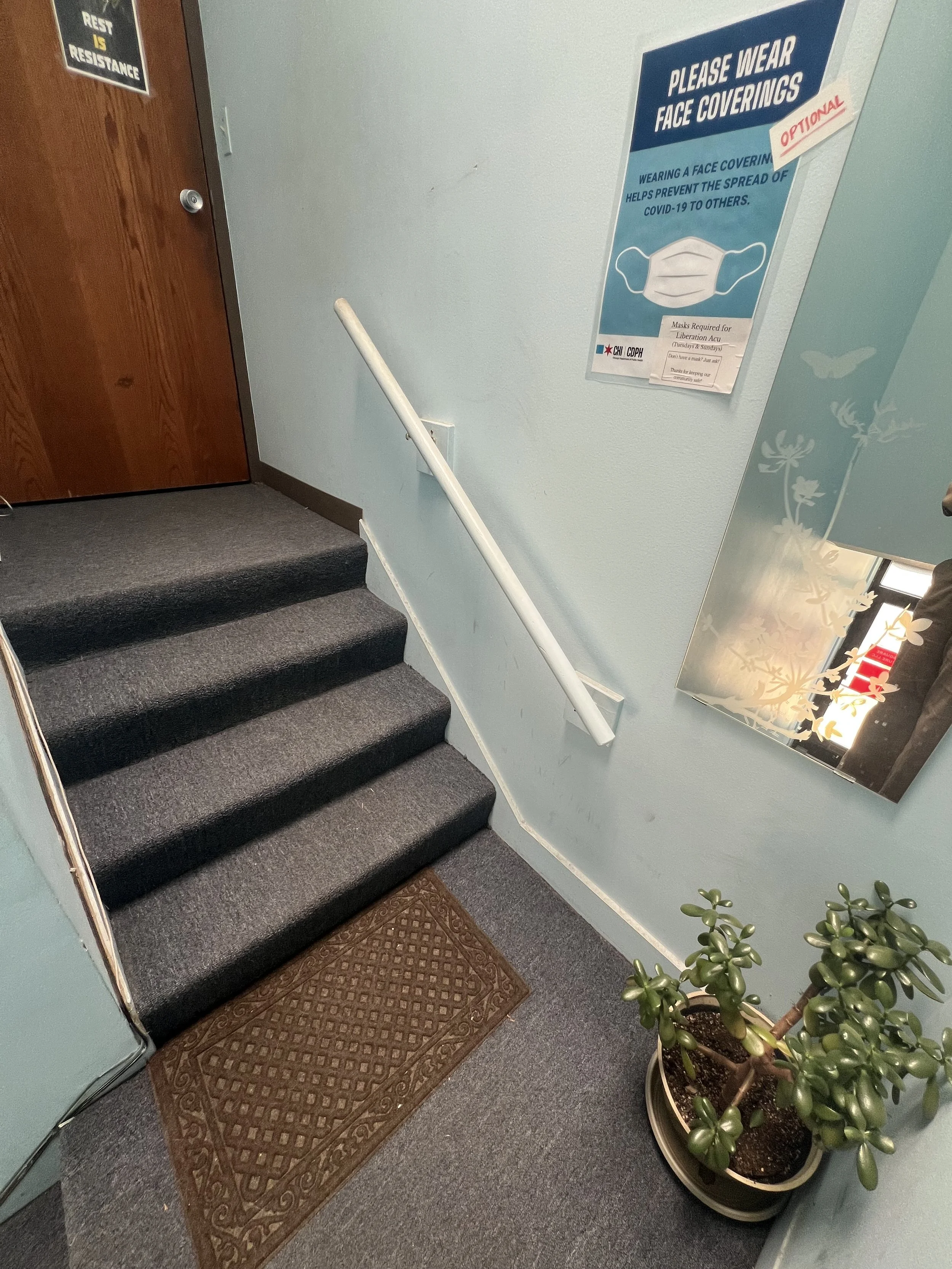 A small platform at the top of the stairs with a mirror, plant, & sign stating that masks are required for Liberation Acu. There are 4 more carpeted stairs on the left, with a hand rail on the right-hand side. The door pushes open into the clinic.