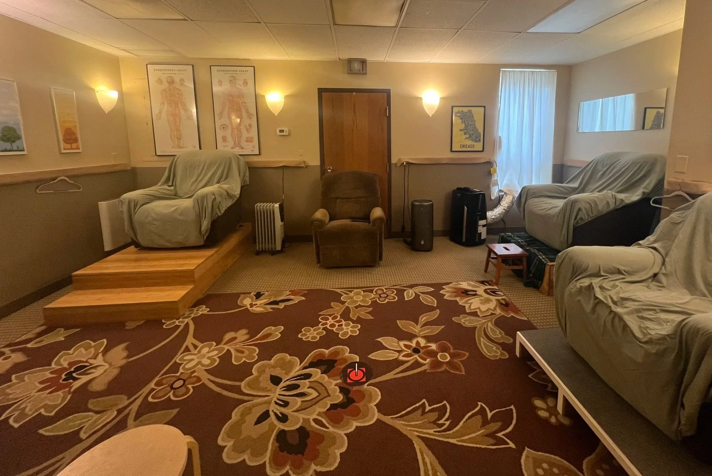 The main treatment room featuring warm lighting, one small window, 4 cushioned reclining chairs sitting on various hand-made wooden platforms, a large floral brown and maroon area rug, an air purifier, AC unit, space heater, and framed posters.