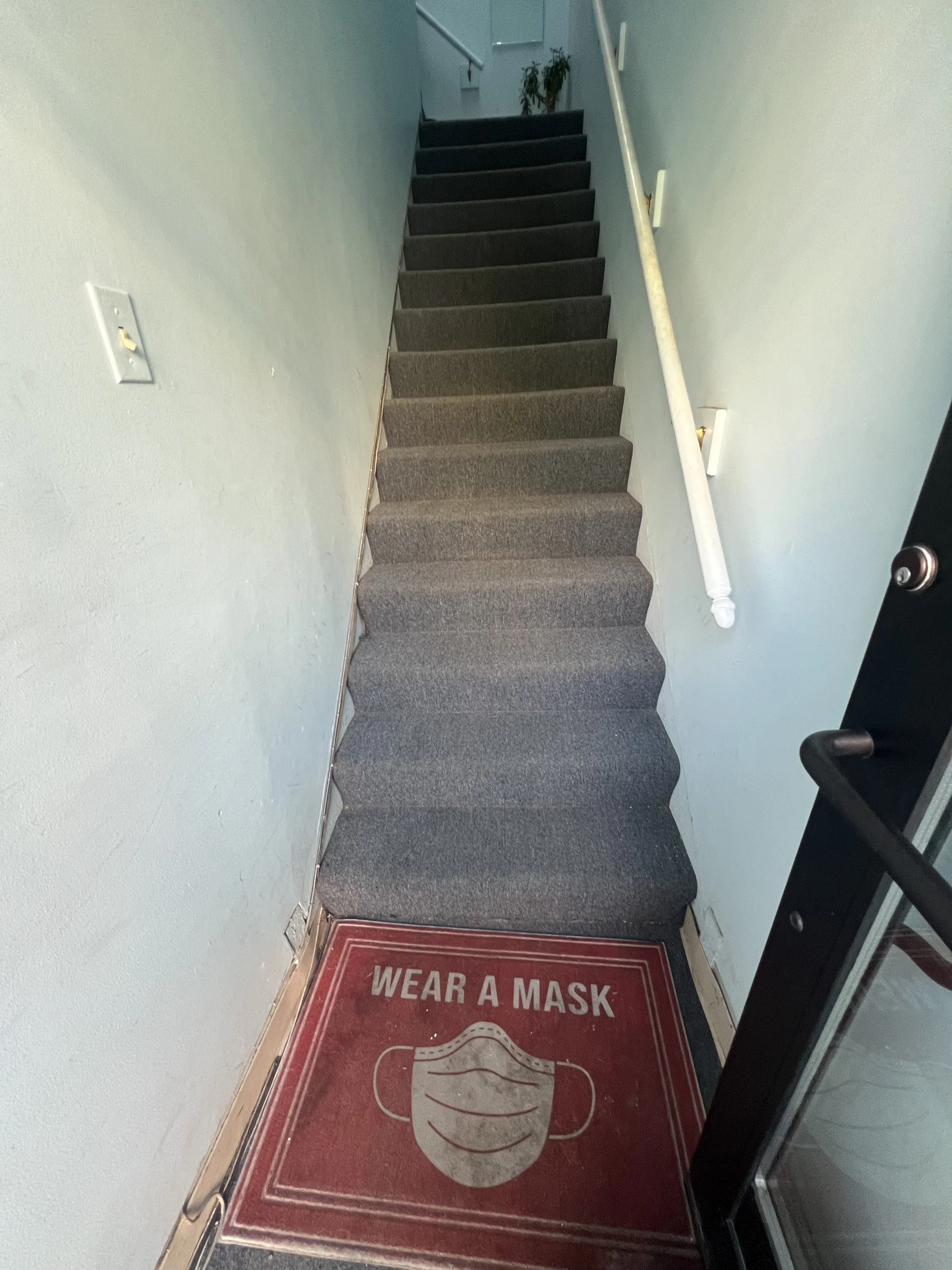 The street-facing door pushes open into the entryway that has a red door mat stating "Wear A Mask". There are 15 stairs with grey carpeting and a hand rail on the right-hand side, leading to a small platform at the top.