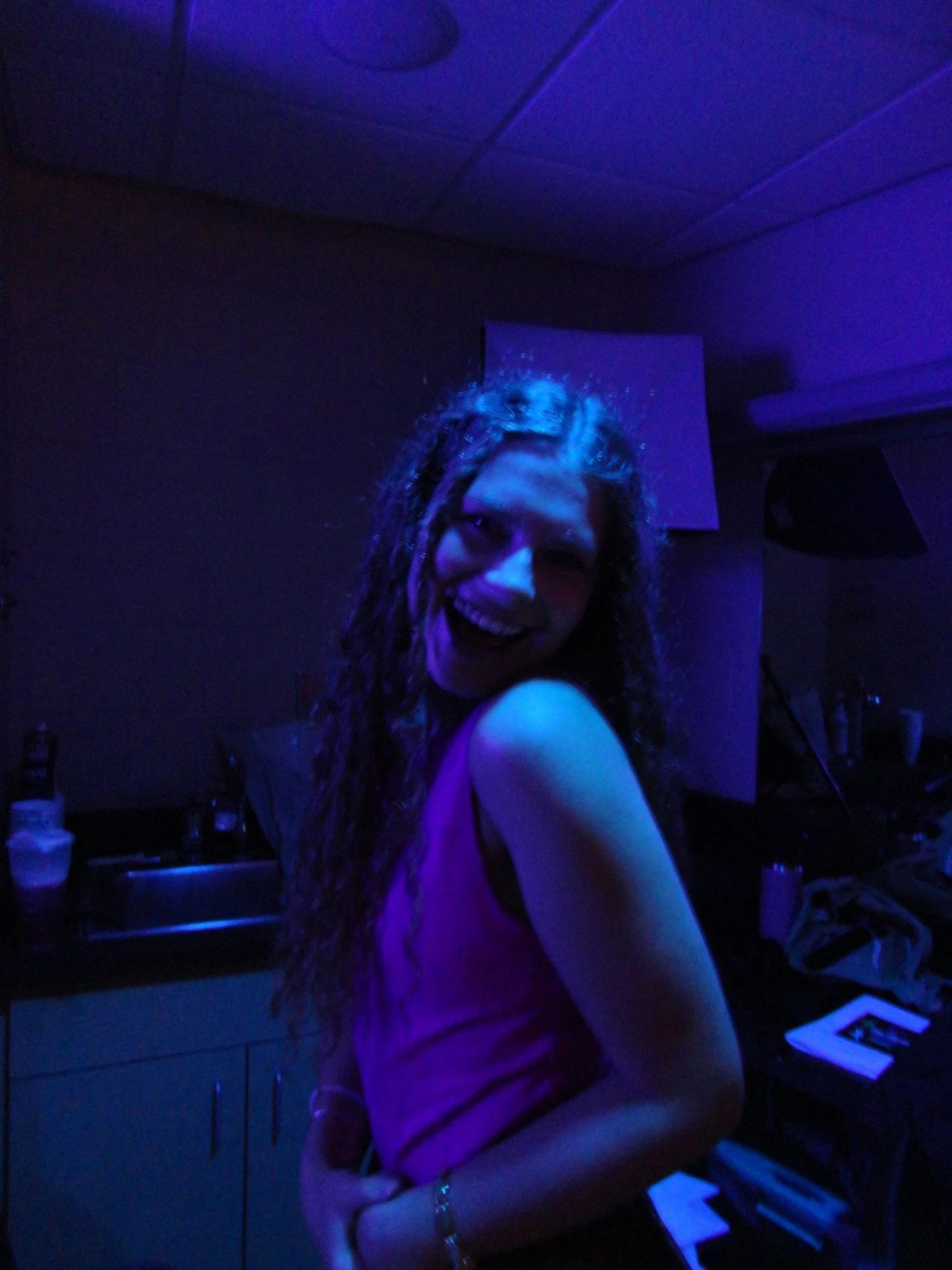 A woman with long curly hair smiling in a dark room illuminated by blue purple lighting. She is wearing a sleeveless top and a bracelet, with some objects on the kitchen counter and a speaker visible in the background.