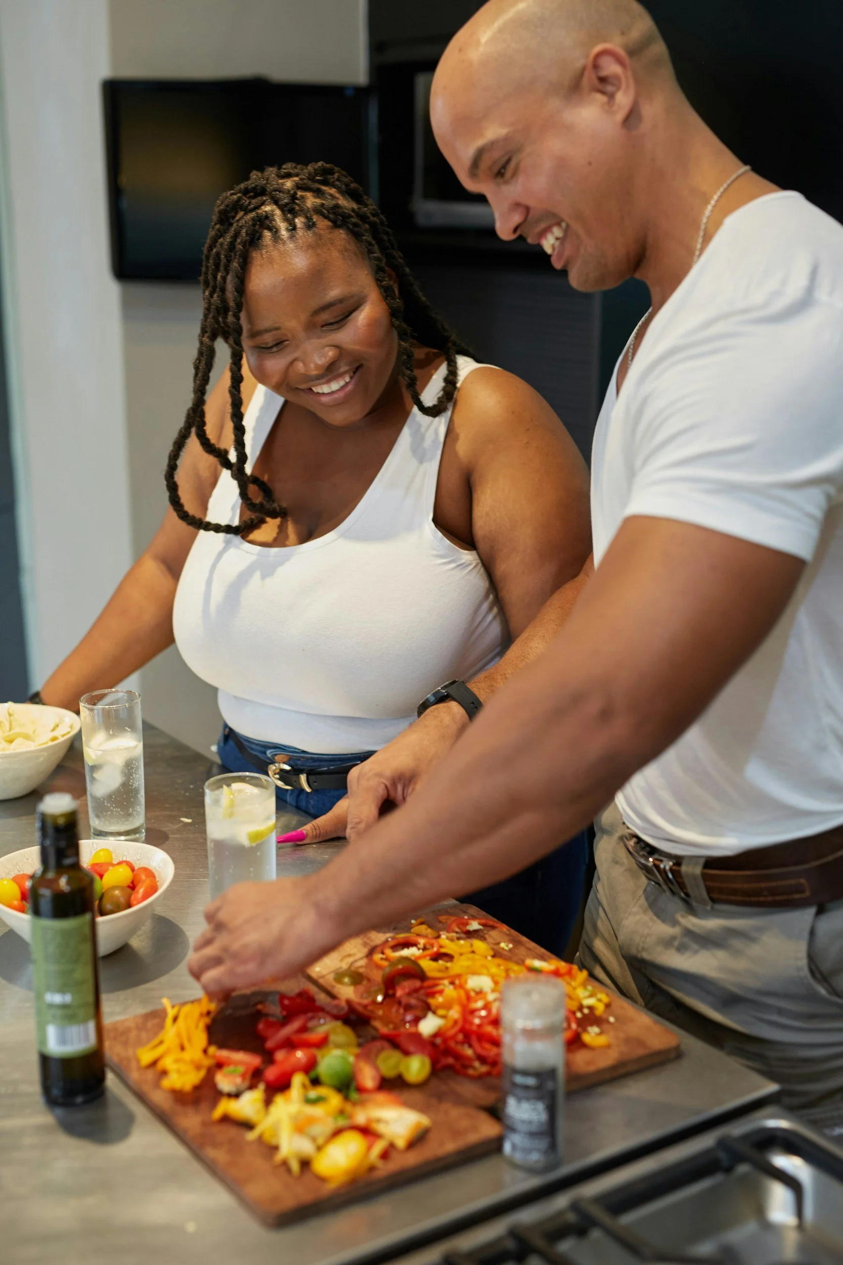 A man and woman preparing homemade pizza together in a kitchen, surrounded by ingredients such as chopped vegetables and popping glasses of water.
