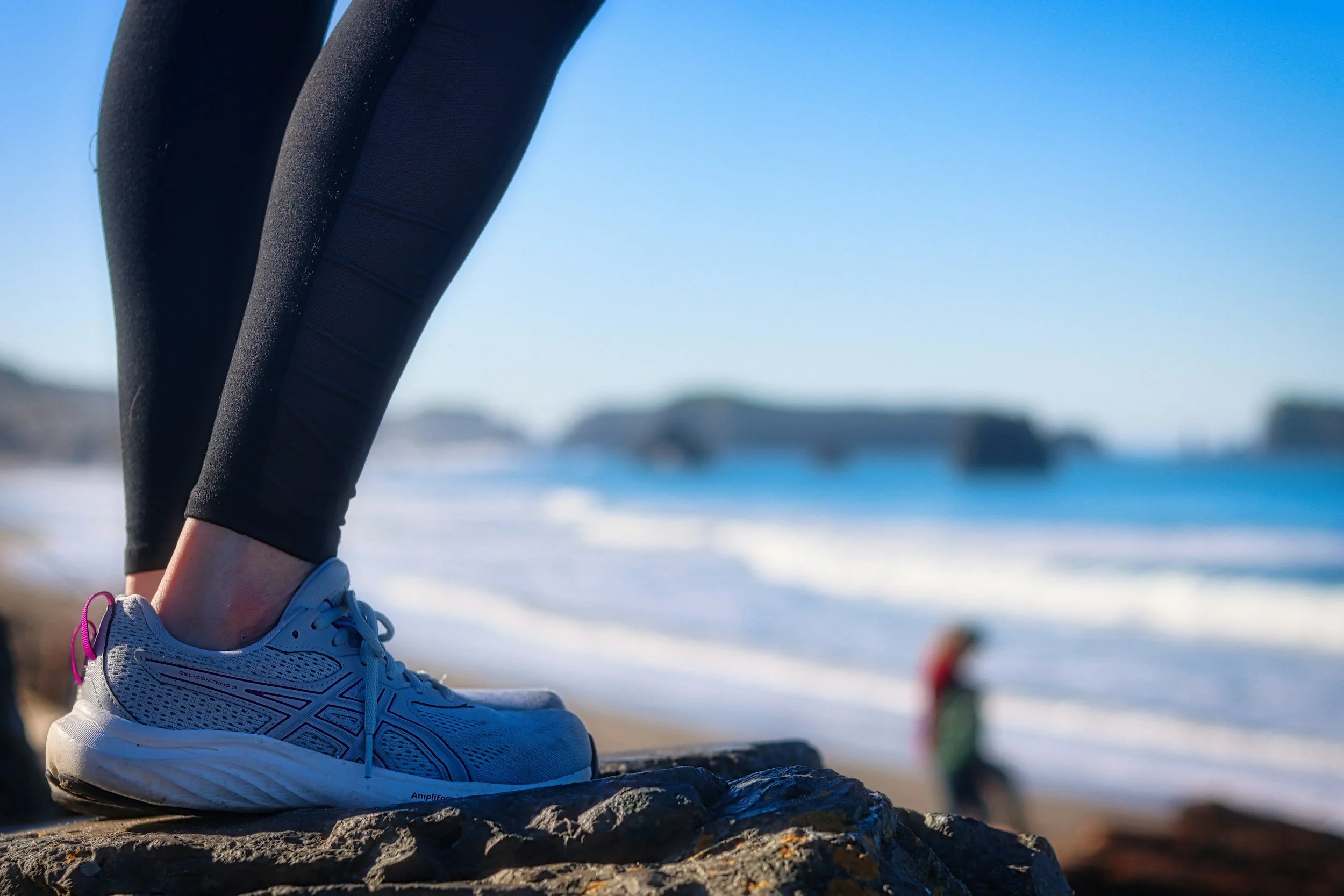 Close-up of a person's foot in a white athletic shoe resting on a rock at the beach, with the ocean and sky in the background.