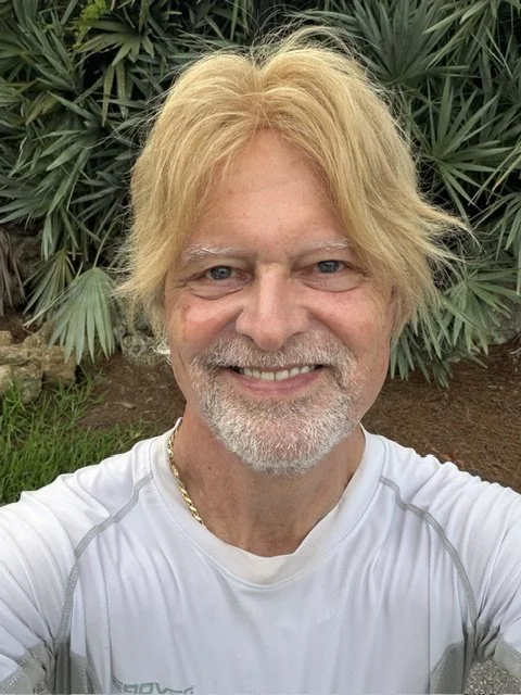A smiling man with blond hair, blue eyes, and a beard, wearing a white athletic shirt, standing outdoors in front of green plants.