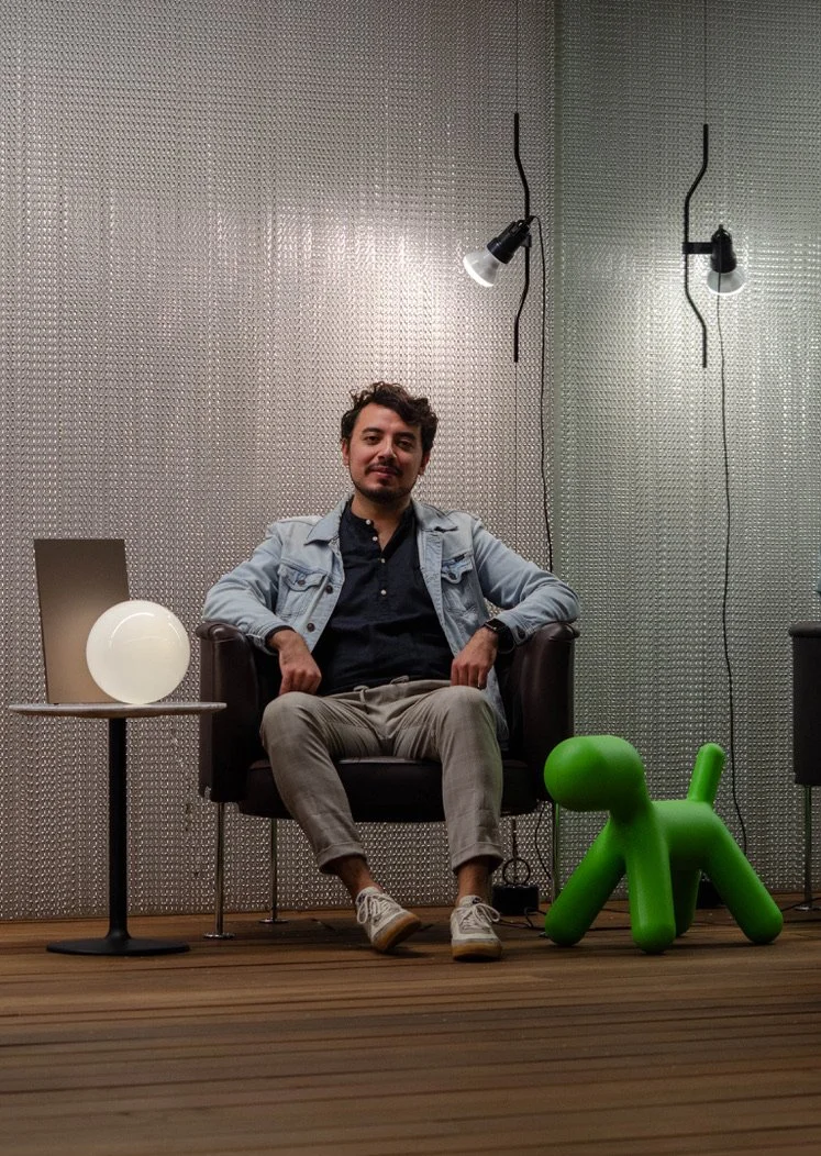 Hombre sentado en una silla de color negro, con fondo de pared metálica, mirando hacia adelante, con lámparas en la pared y una lámpara en una mesa a la izquierda, y un perro de plástico verde en el suelo a la derecha.