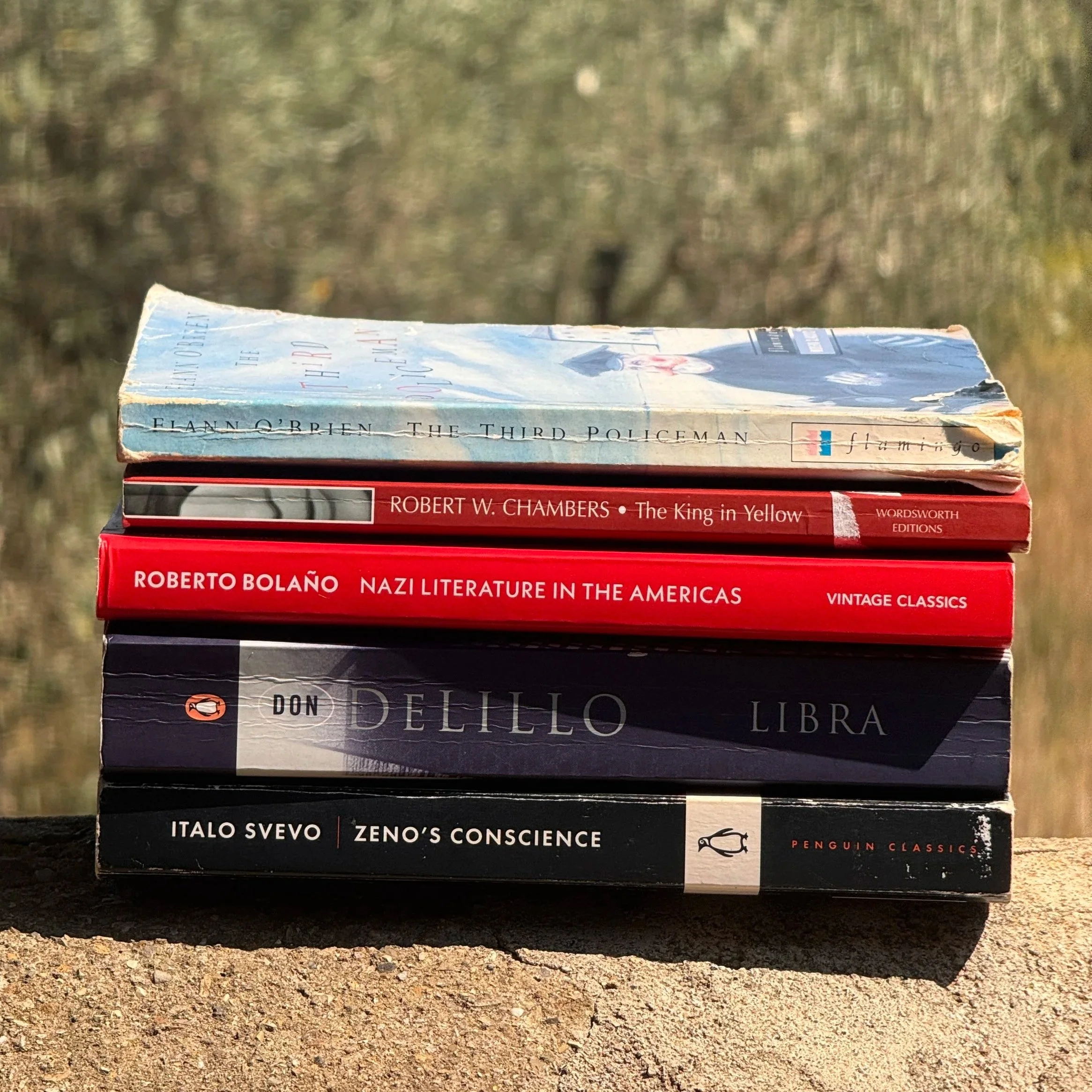 A stack of four books placed on a gravel ground with blurred trees in the background. The titles include 'The Third Policeman' by Flann O'Brien, 'Nazi Literature in the Americas' by Roberto Bolaño, and 'Zeno's Conscience' by Italo Svevo.