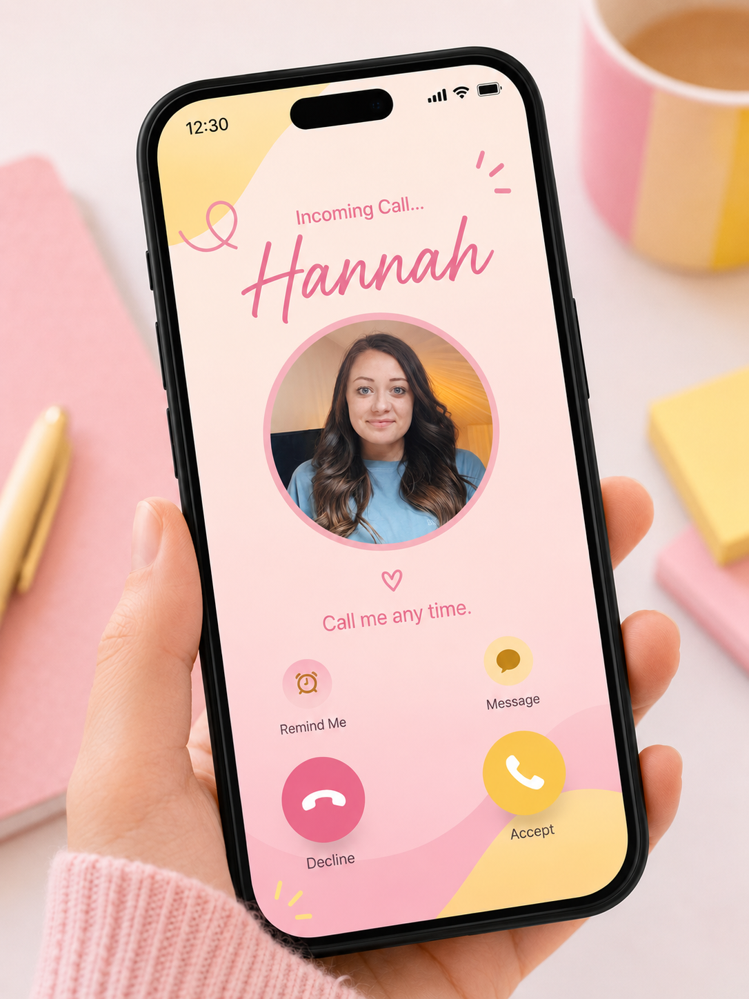 Smartphone screen showing an incoming call from Hannah with options to accept, decline, send message, or set a reminder, held by a person's hand over a pink background with notebooks and cups.