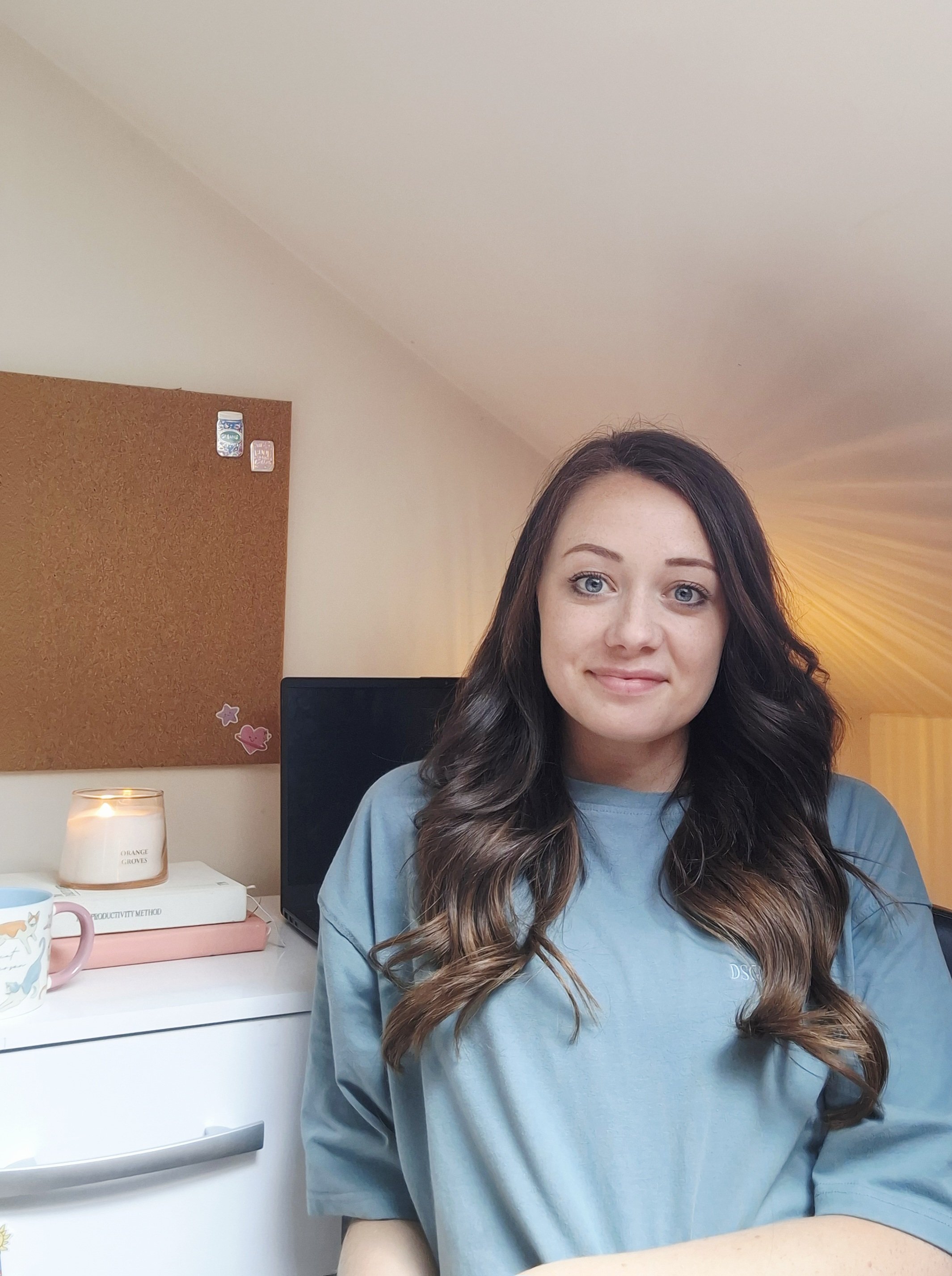 A woman with long dark wavy hair and blue eyes is sitting in a room, wearing a blue shirt. There is a corkboard with some decorations, a candle, a pink book, and a coffee mug on a white desk beside her. A computer monitor is visible in the background, and the room has a warm, glowing light from a lamp.