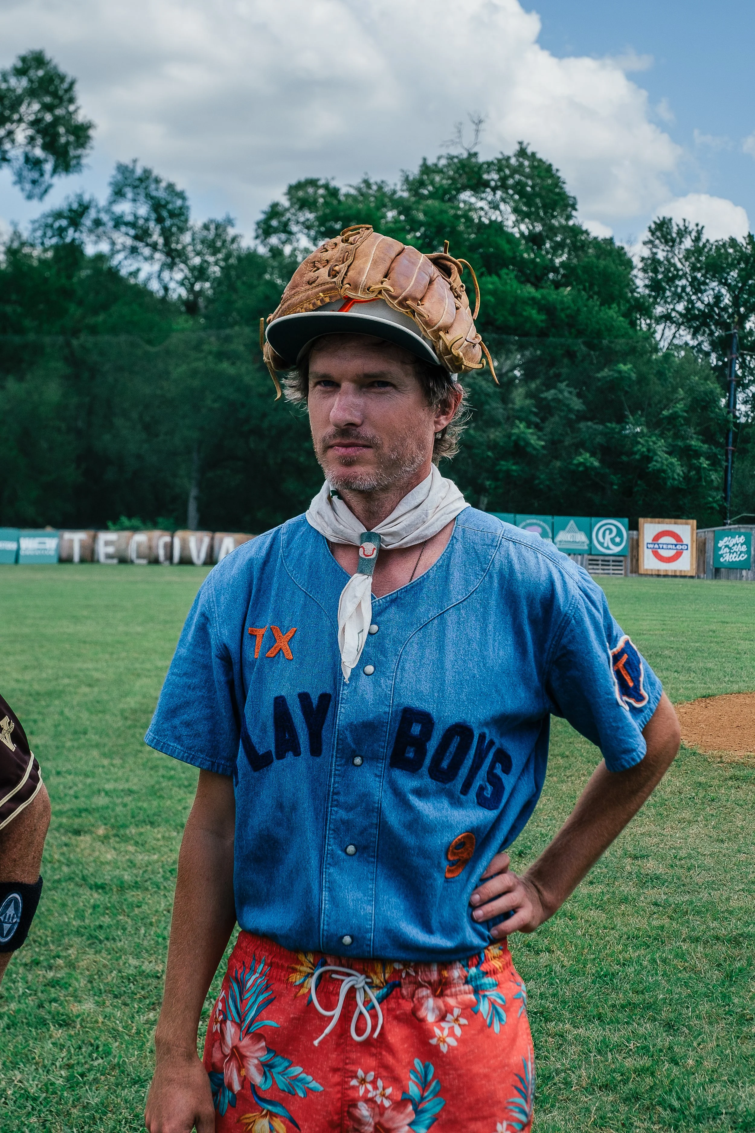 A man standing outdoors on a grassy field, wearing a vintage blue baseball jersey.