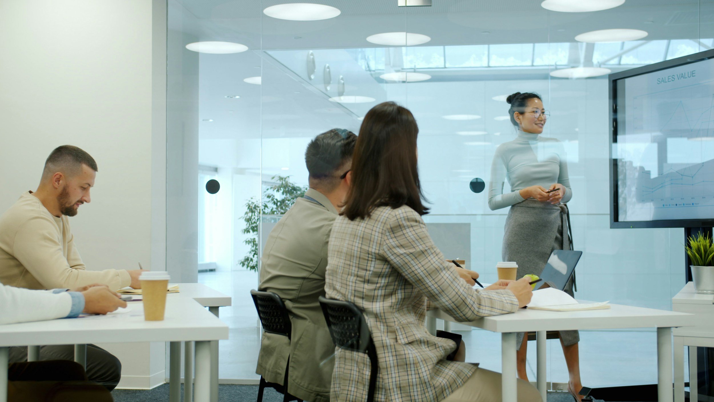 A woman standing near a large screen displaying sales data, giving a presentation to four seated people in a modern conference room.