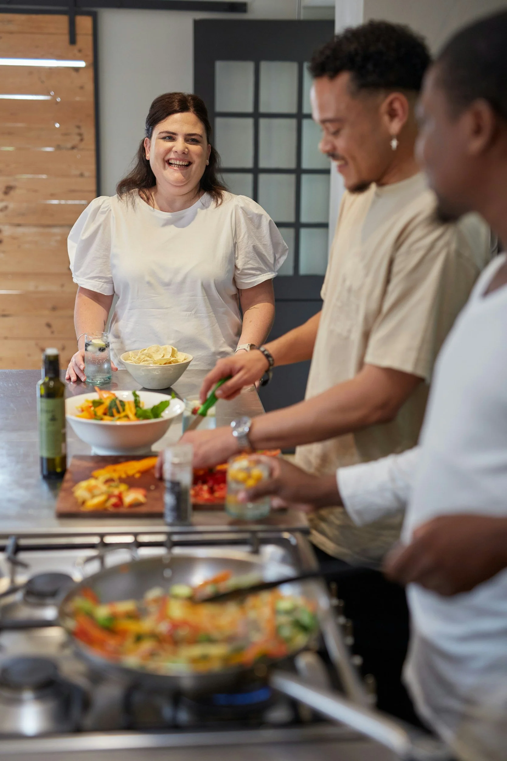 A group of people cooking and enjoying a meal in a modern kitchen, with one woman smiling at the camera.
