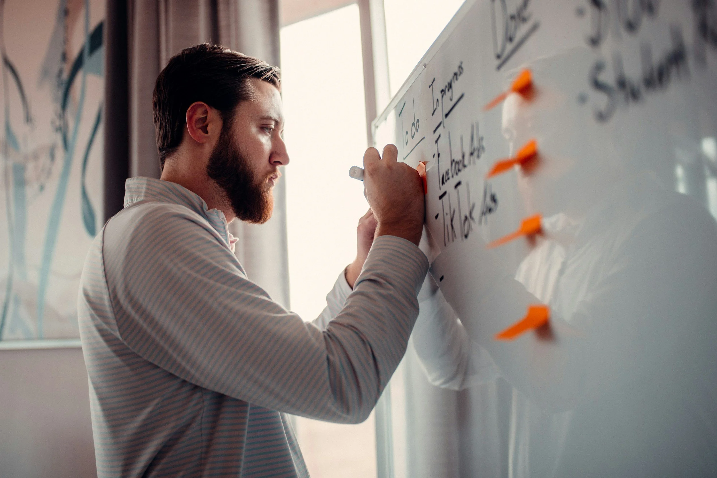 A man with brown hair and a beard writing on a whiteboard with orange sticky notes attached to it, in a bright room with large windows.