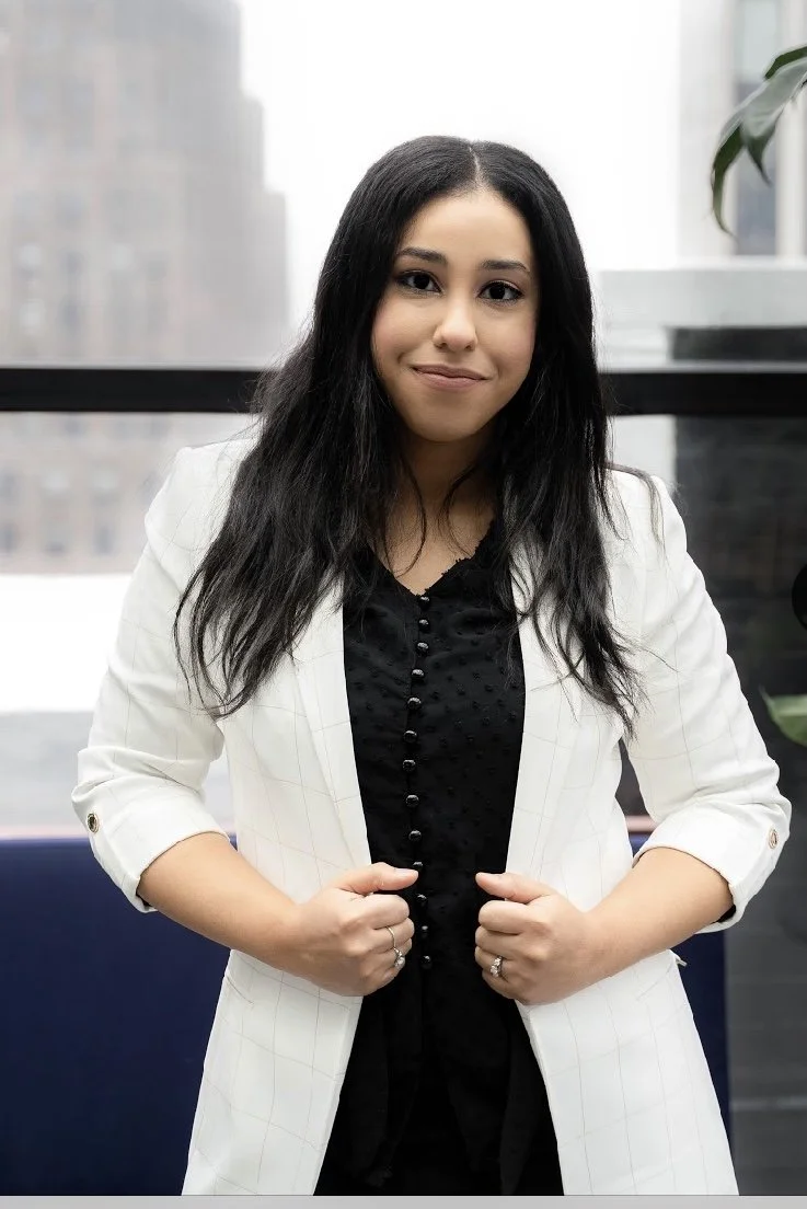 A woman with long black hair, wearing a white blazer over a black shirt, standing indoors in front of a window with a cityscape in the background.