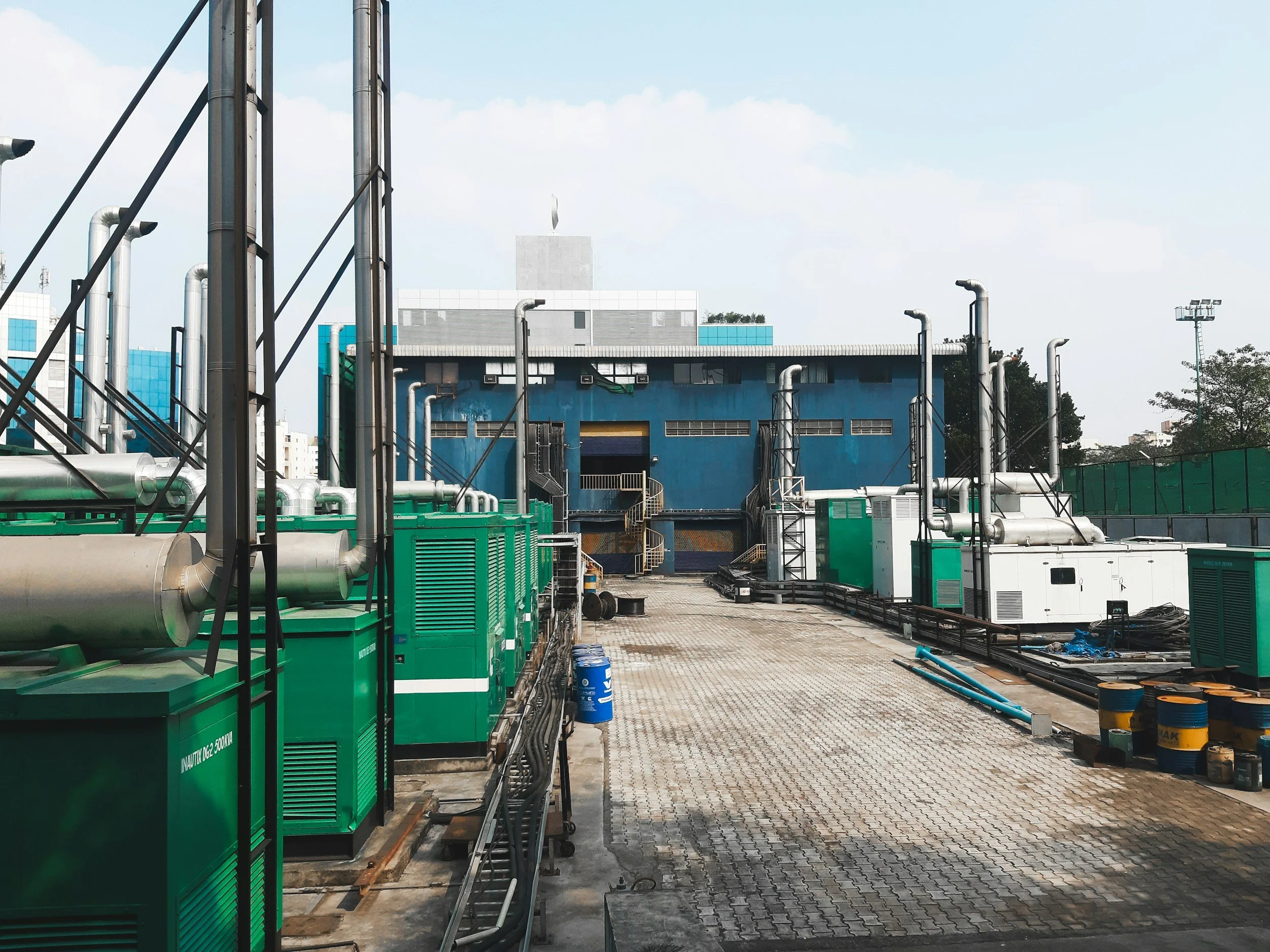 Industrial facility with numerous green generators, metal pipes, and structural beams under a partly cloudy sky.
