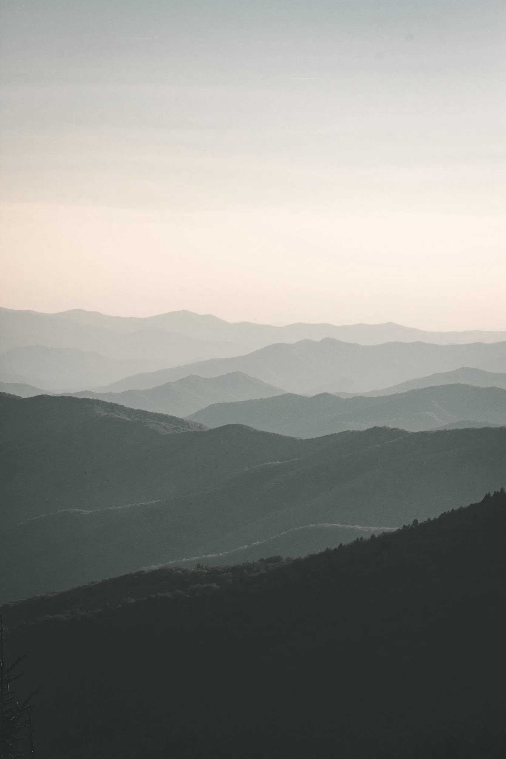 Hazy mountain landscape with multiple layers of mountain ridges receding into the distance under a pale sky.