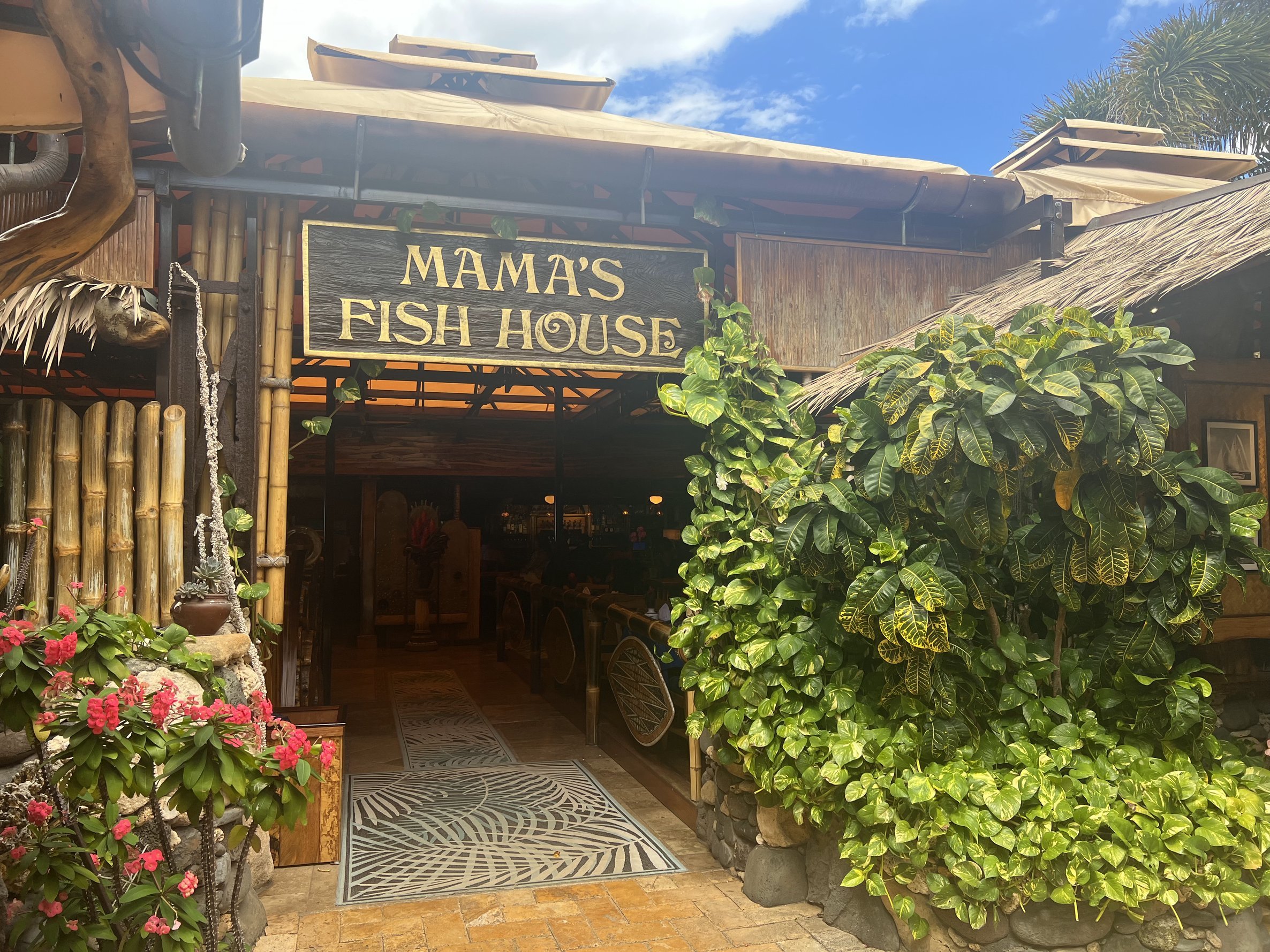 Entrance to Mama's Fish House with tropical plants, bamboo fencing, and a thatched roof, under a blue sky with clouds.