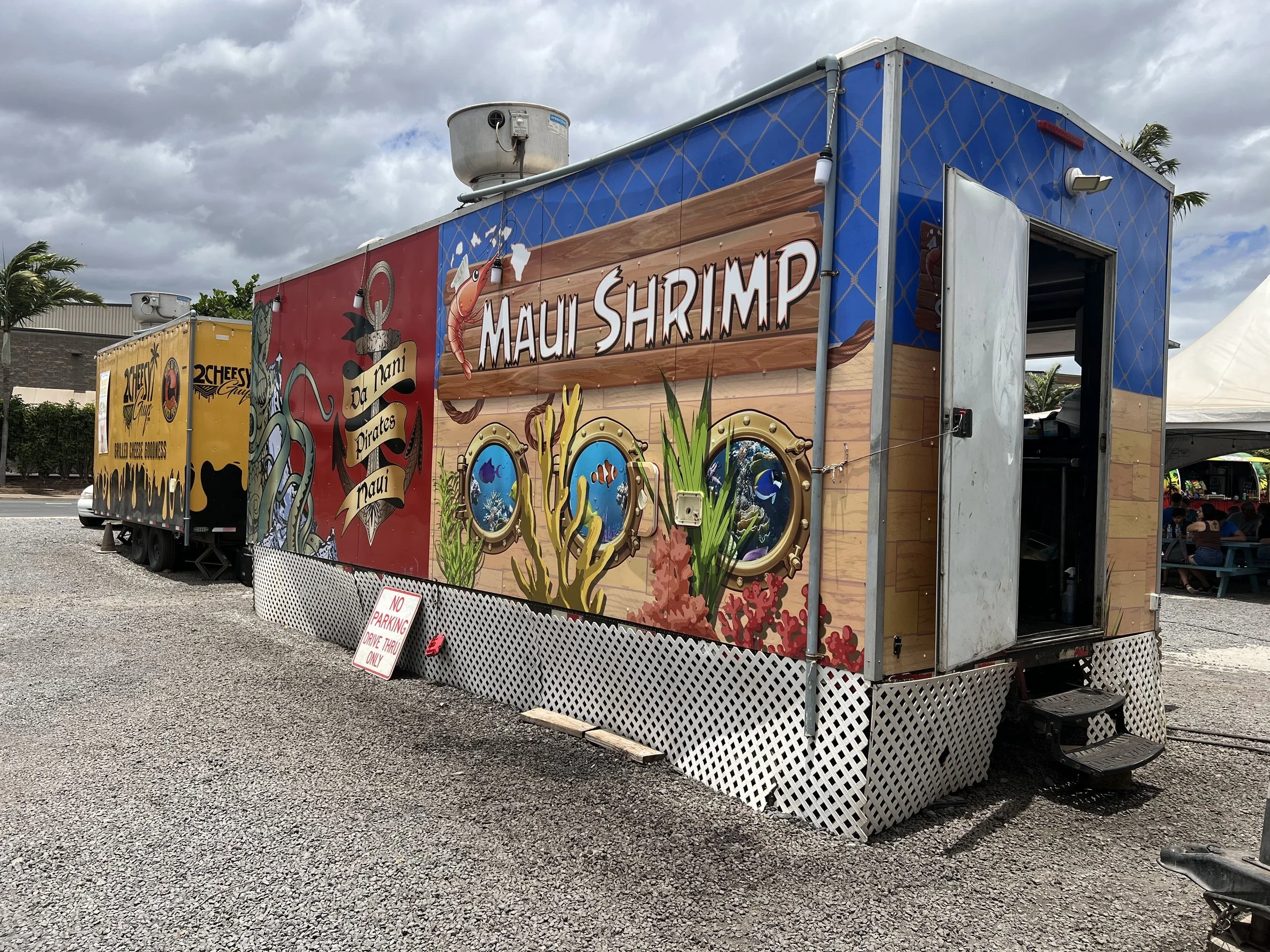Colorful food truck with a sign that reads "Maui Shrimp" parked outdoors under a cloudy sky, with another yellow food truck behind it and people visible in the background.
