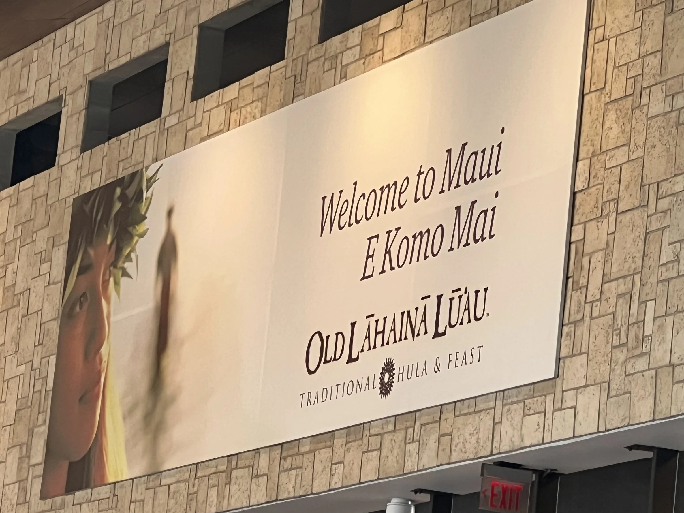 A tiled brick wall with a large white sign that has writing promoting a traditional Hawaiian feast called 'E Komo Mai'. The sign also features a colorful flower arrangement and a hand holding a yellow flower.