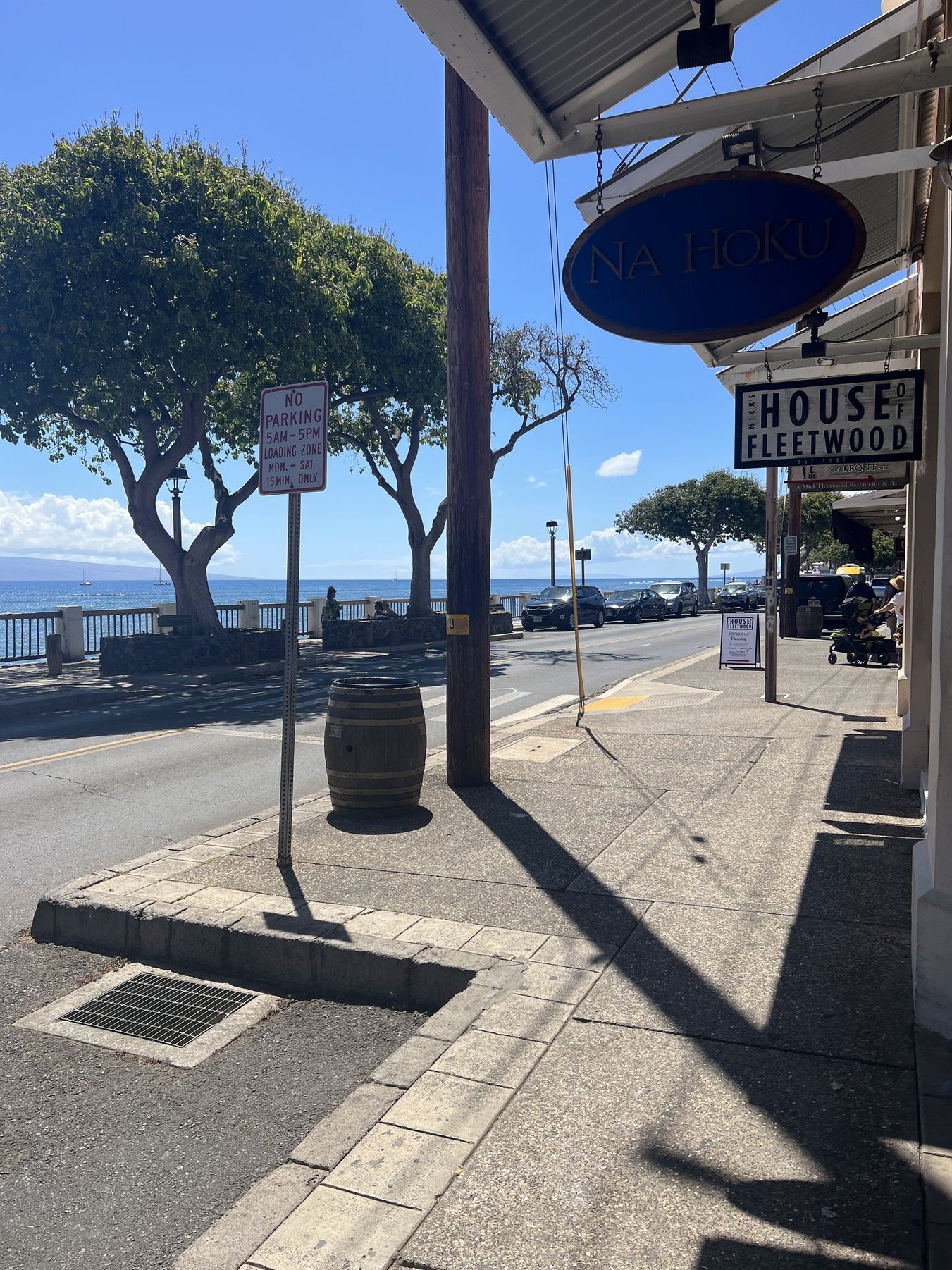 A sidewalk by the waterfront with cars parked along the street, trees, and a sign that says 'No Parking 5am-5pm Loading Zone Mon.-Sat. 15 Min. Only'. There are some trees, a street lamp, and a view of the ocean with a sailboat in the background. The sky is clear and blue.