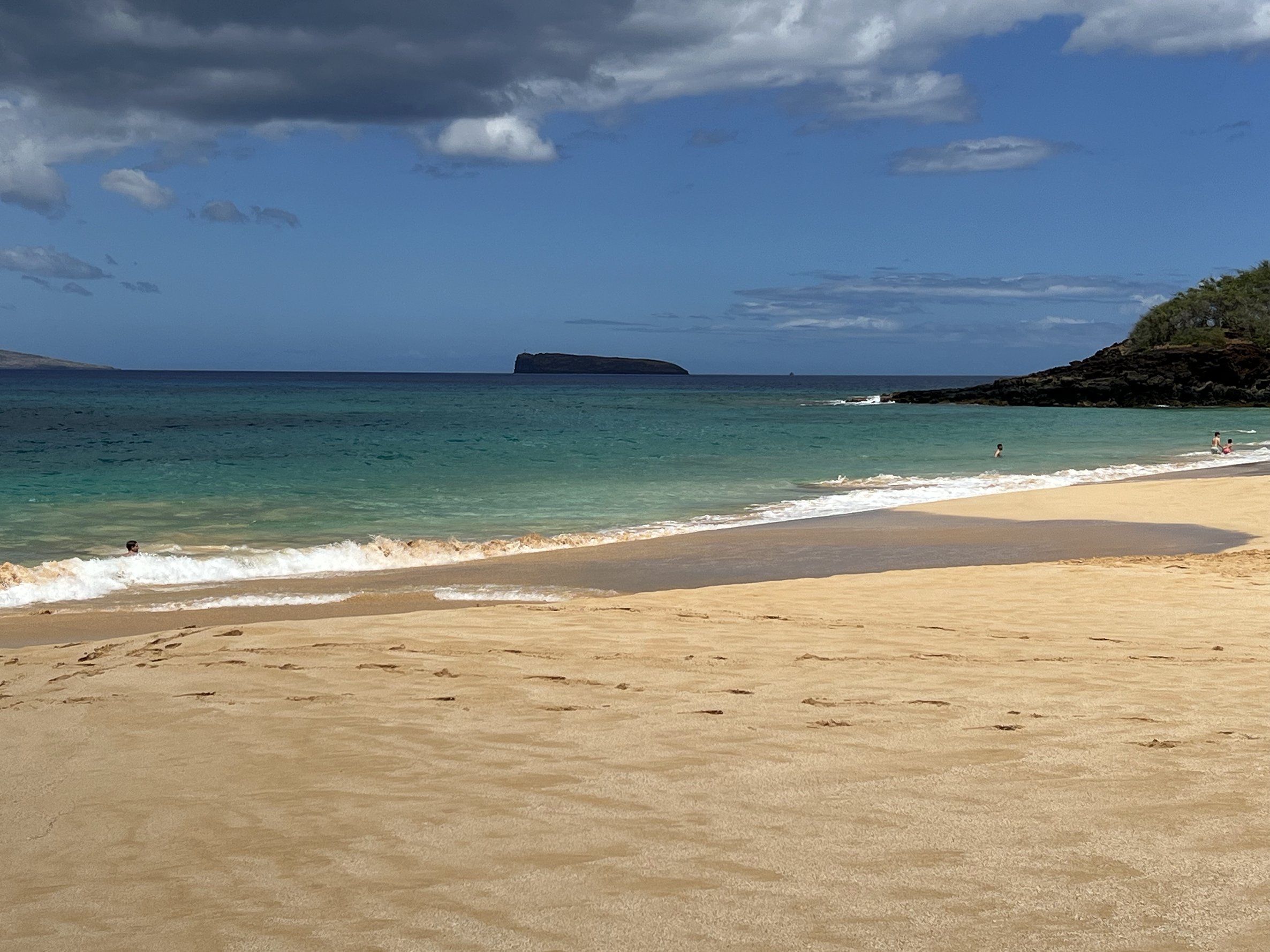 A sandy beach with a few people near the shoreline, calm turquoise water, a rocky headland on the right, and a distant landmass on the left under a partly cloudy sky.